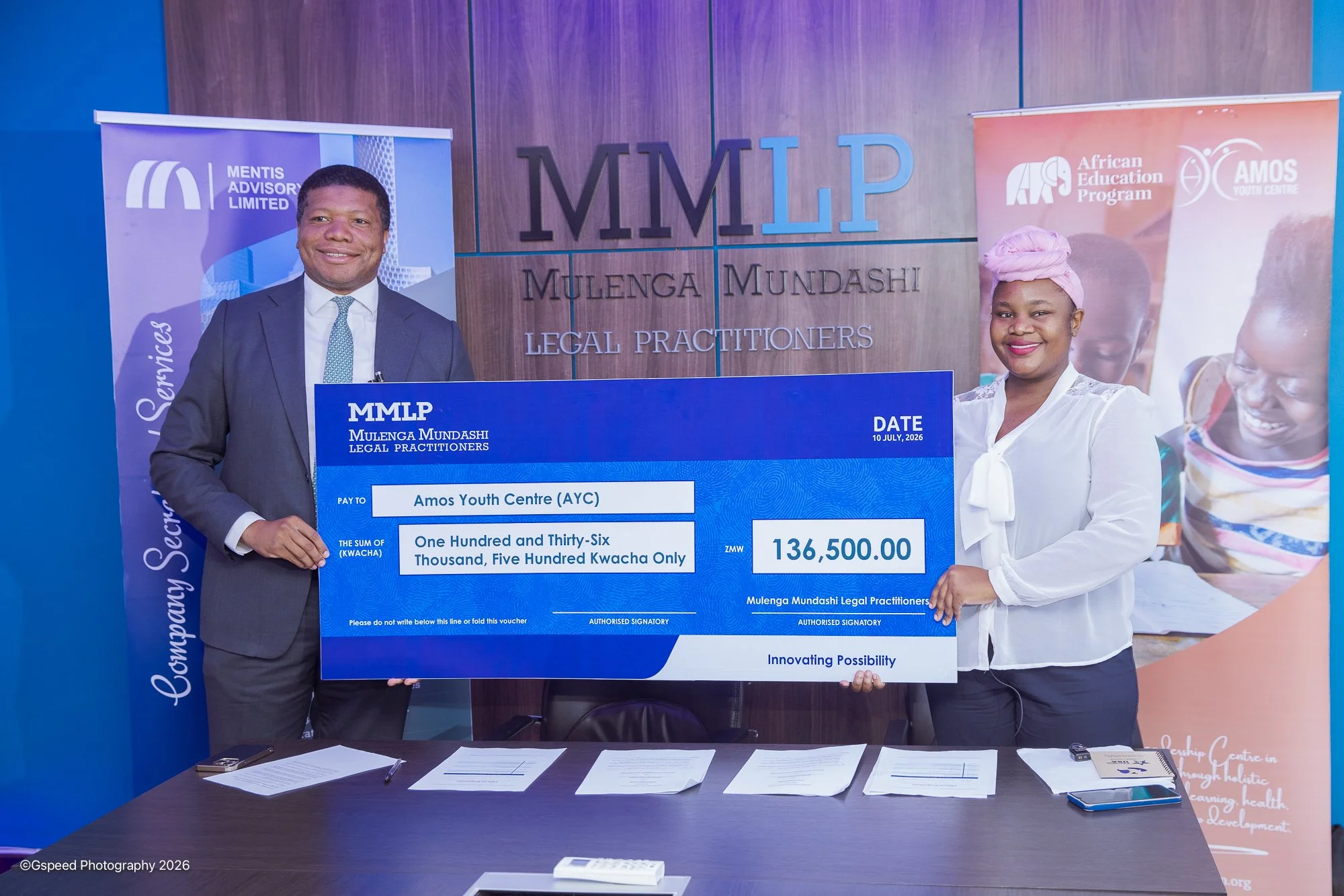 Two individuals, a man in a suit and a woman in a white blouse with a pink headwrap, holding a large ceremonial check for 136,500 ZMW made out to Amos Youth Centre, at an event at Mulenga Mundashi Legal Practitioners. Behind them are banners and a wa