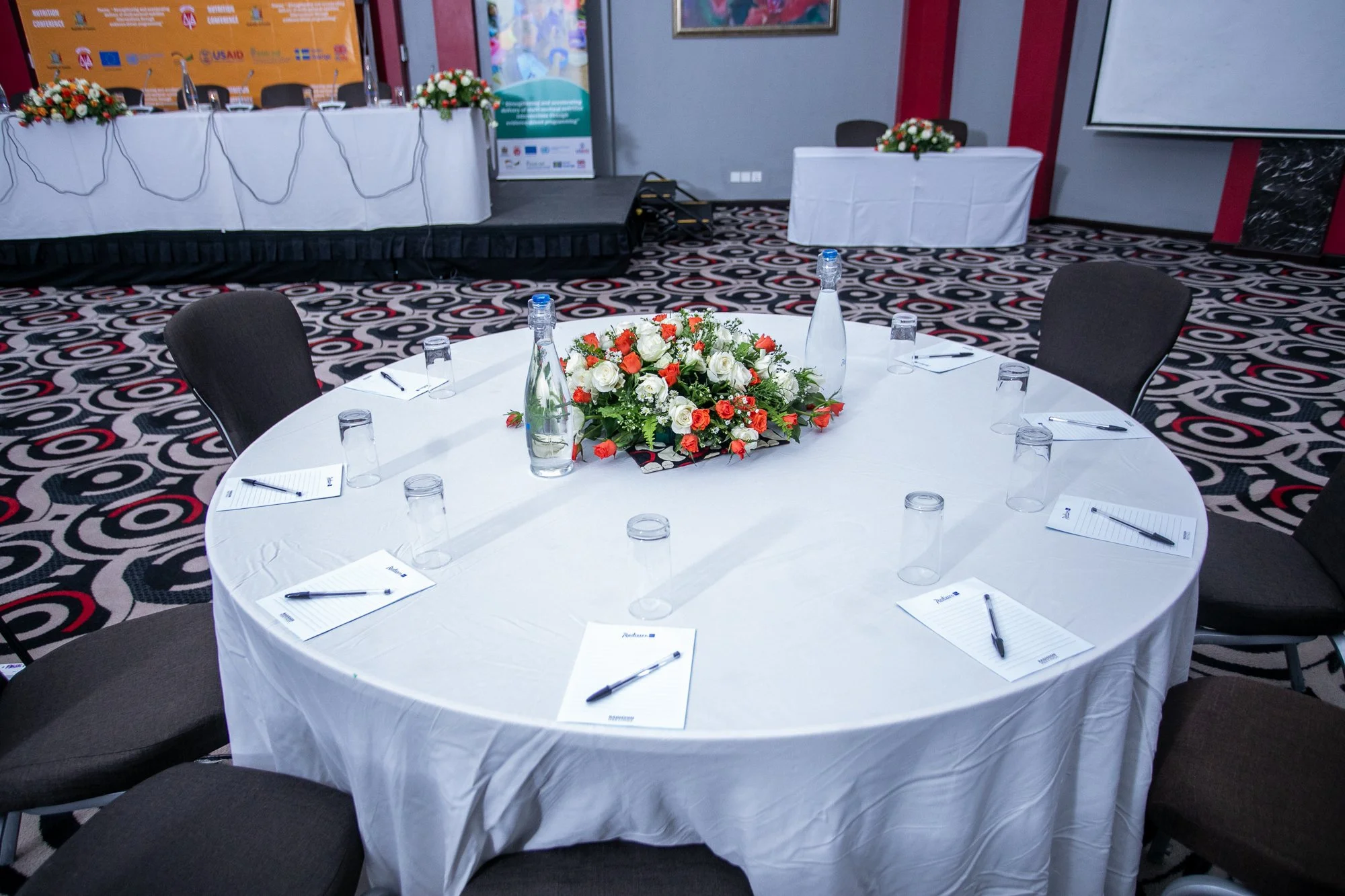 A round conference table covered with a white tablecloth, decorated with a floral centerpiece of white and orange roses. The table is set with notepads, pens, glasses, and bottles of water. In the background, a stage with chairs and a table with flor