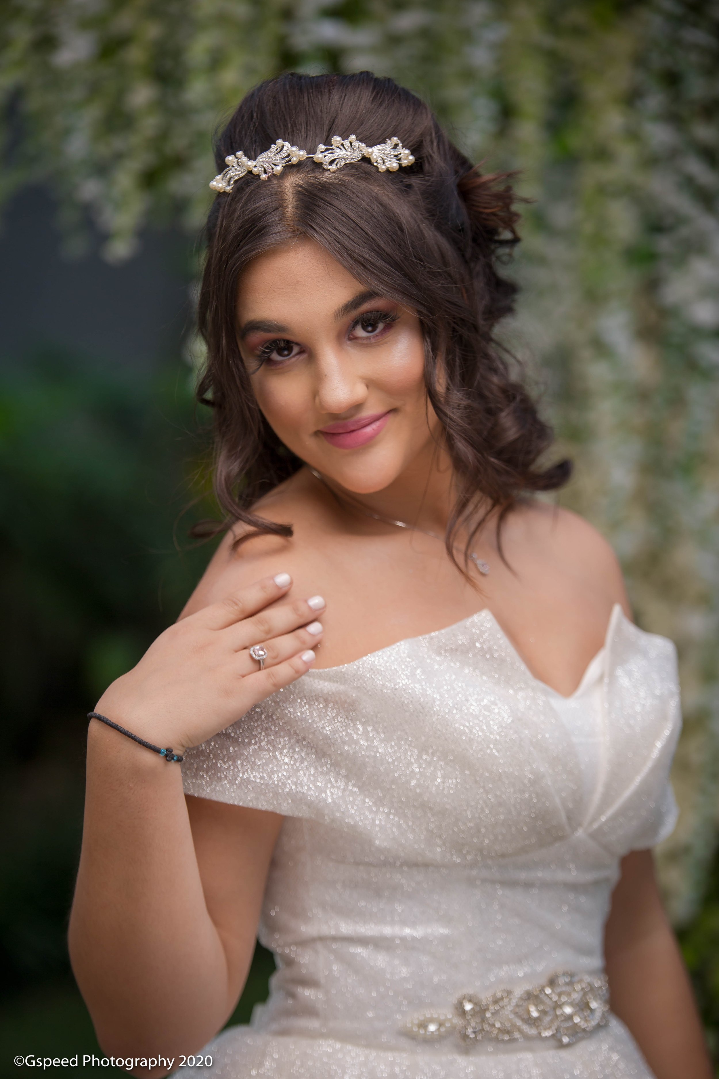 A young woman with brown hair styled in loose curls, wearing a sparkly off-the-shoulder dress and a pearl and jewel headband. She's smiling softly, with makeup on her eyes and lips, in front of a blurred natural background.