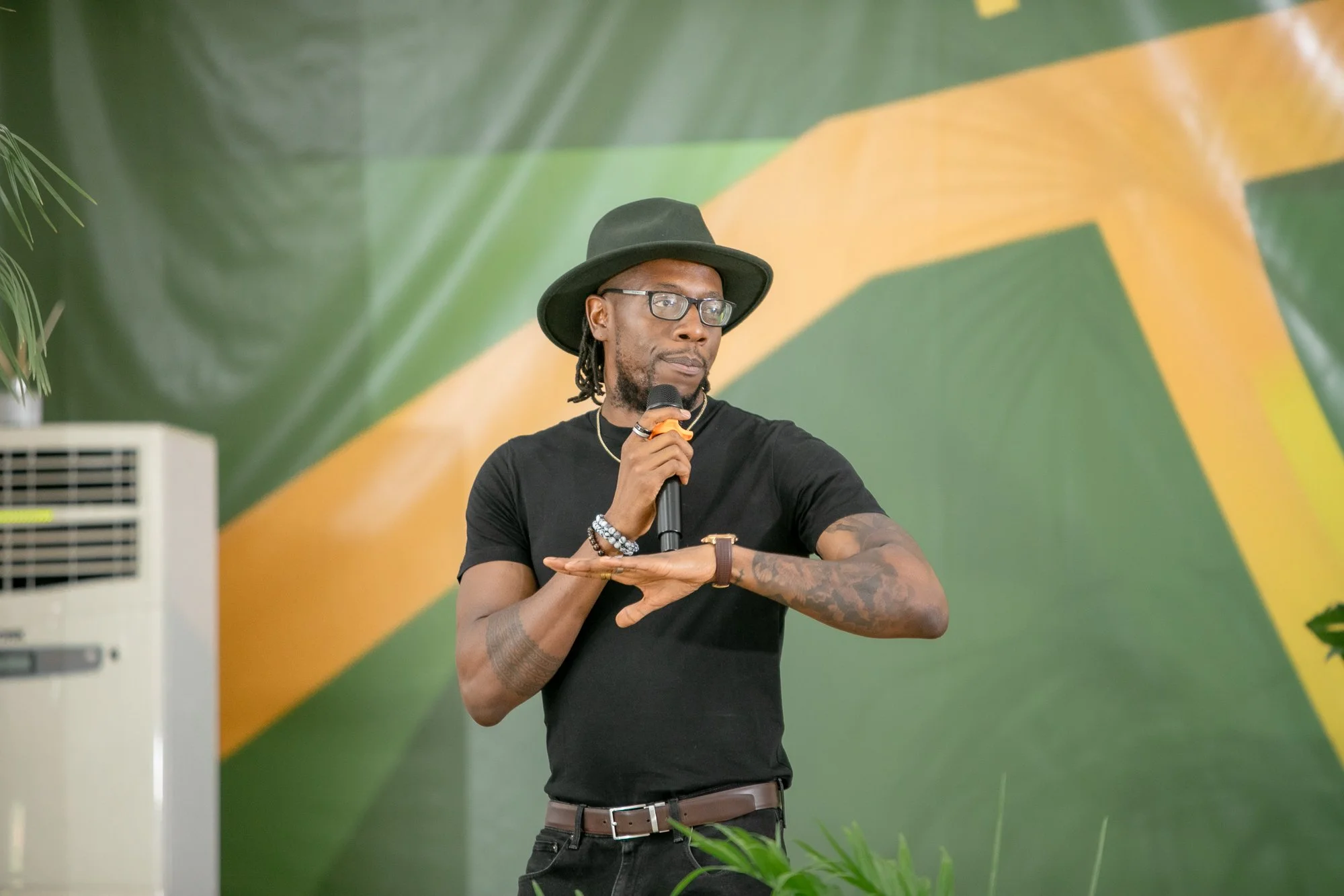 A man with dreadlocks, glasses, and tattoos wearing a black hat and shirt holds a microphone while standing in front of a green and yellow background.