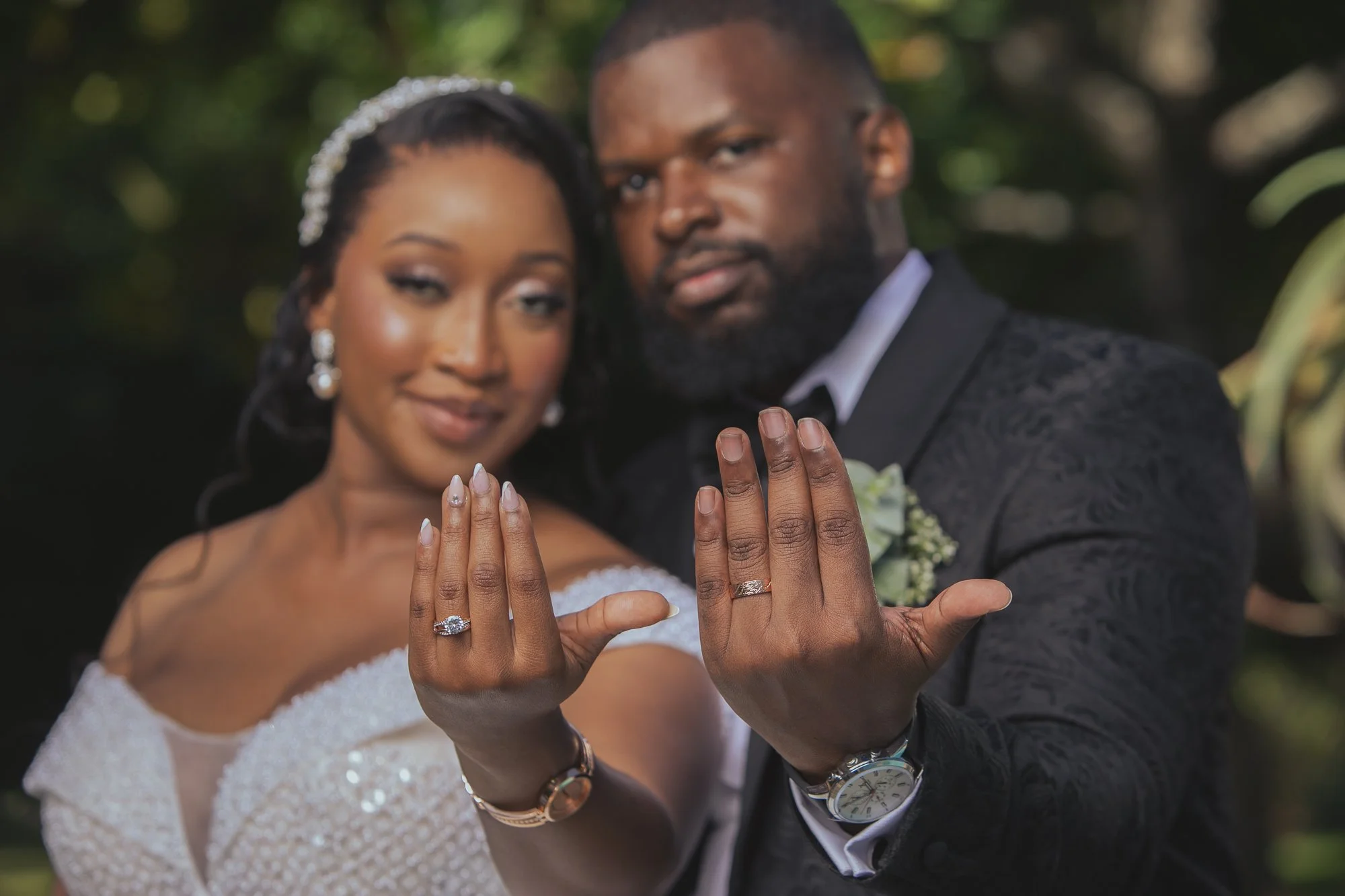 A newlywed couple shows their wedding rings while holding their hands up. The bride wears a white dress and sparkly earrings, and the groom wears a black suit with a watch and boutonniere. They are outdoors with blurred greenery in the background.
