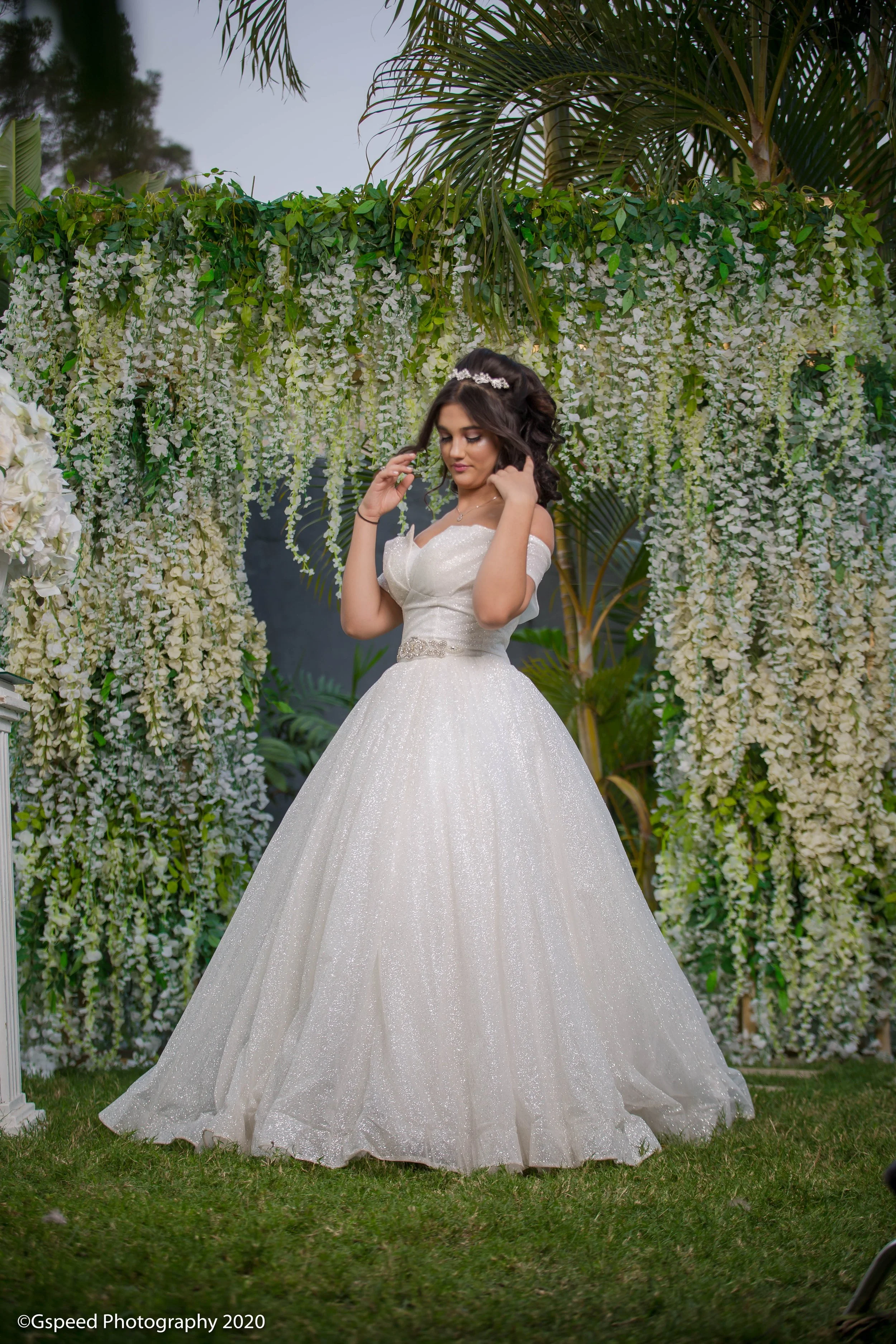 A woman in a white wedding dress standing in front of decorated flower walls with greenery and white hanging flowers, outdoors during daytime.
