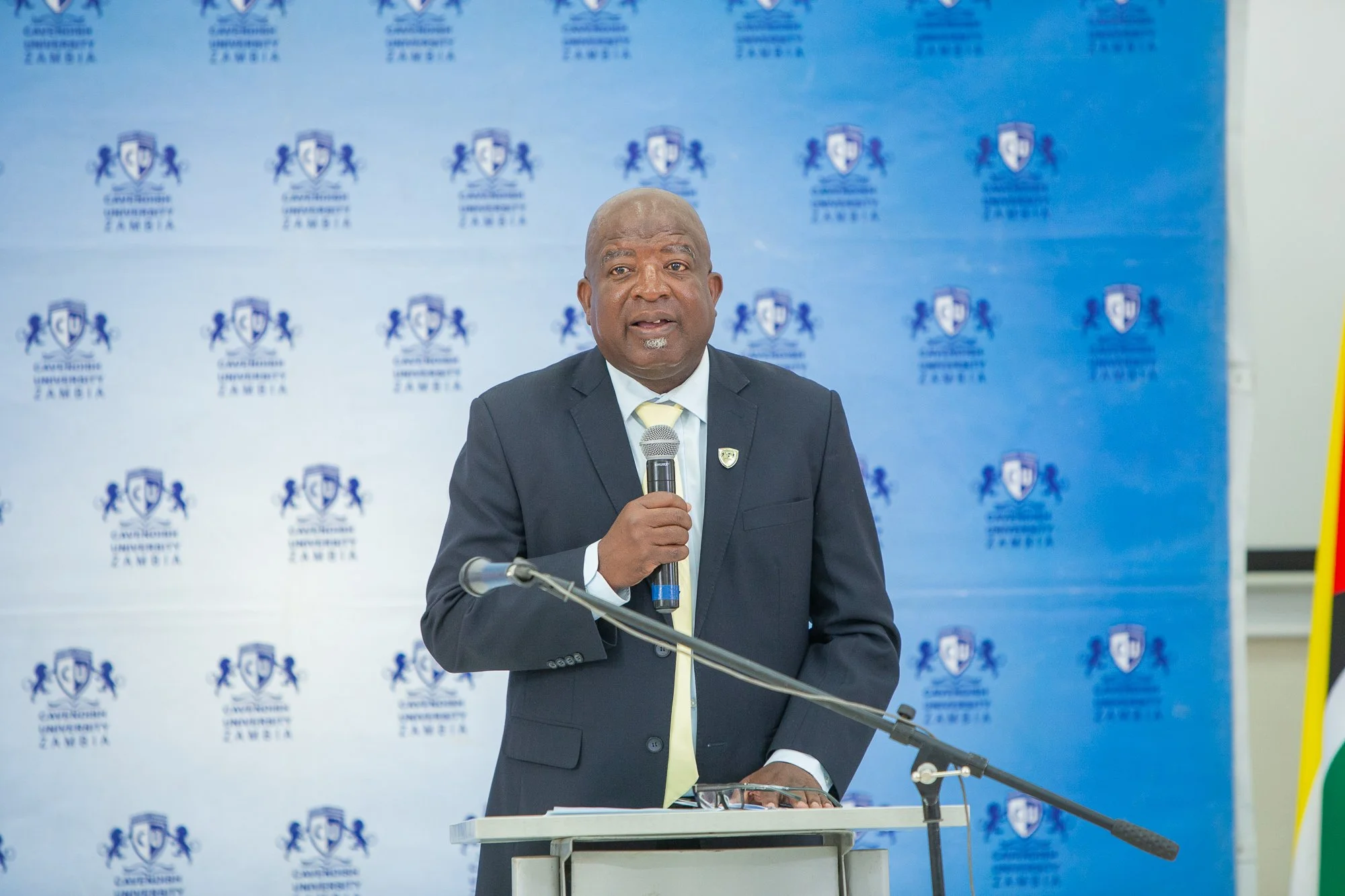A man in a suit and tie speaking into a microphone at a podium during a formal event at Camden University Zambia. The background features a blue banner with the university's logo repeated across it.