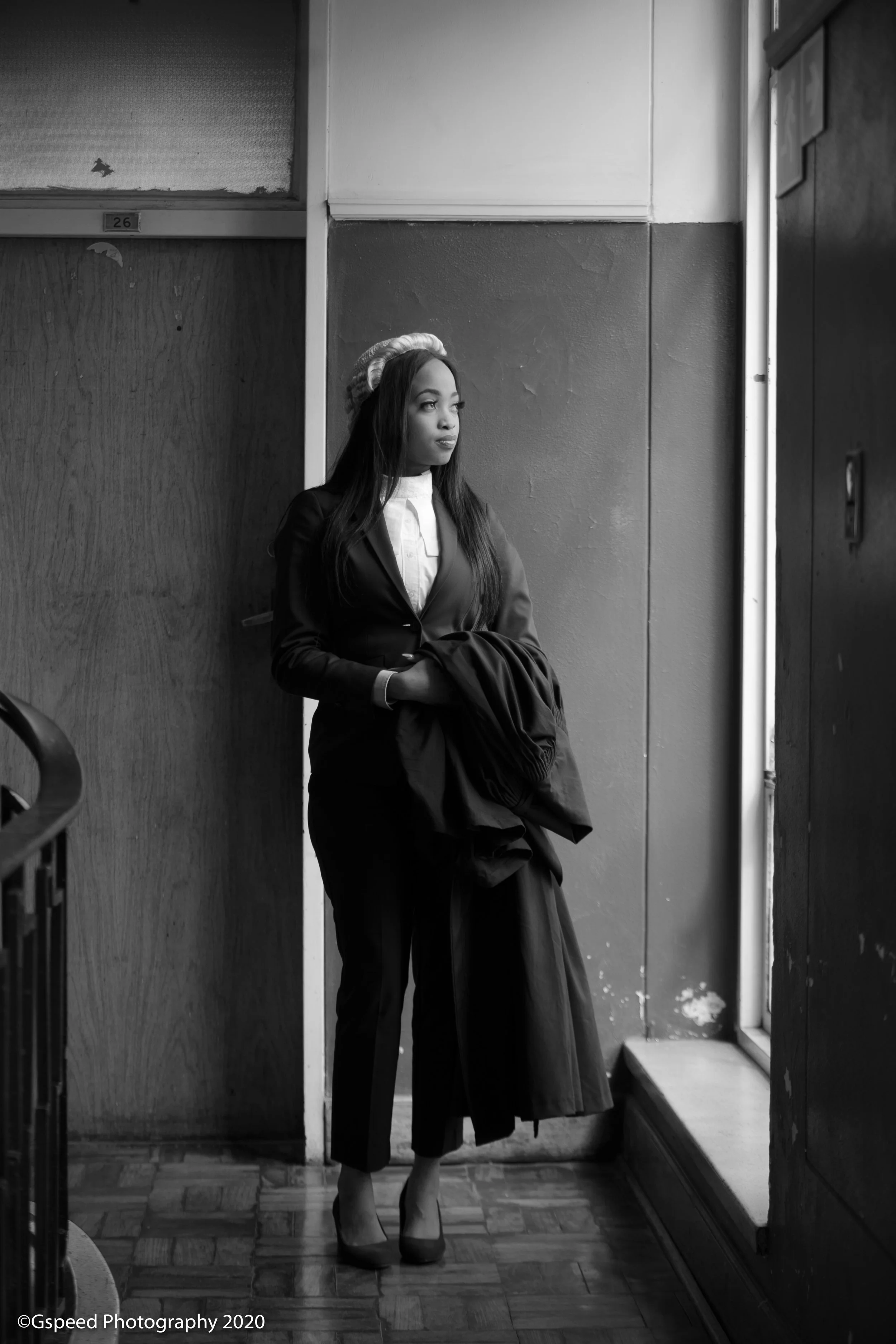 A woman in a suit and high heels stands by a window, looking outside, inside a building with wood-paneled walls and tiled floor.