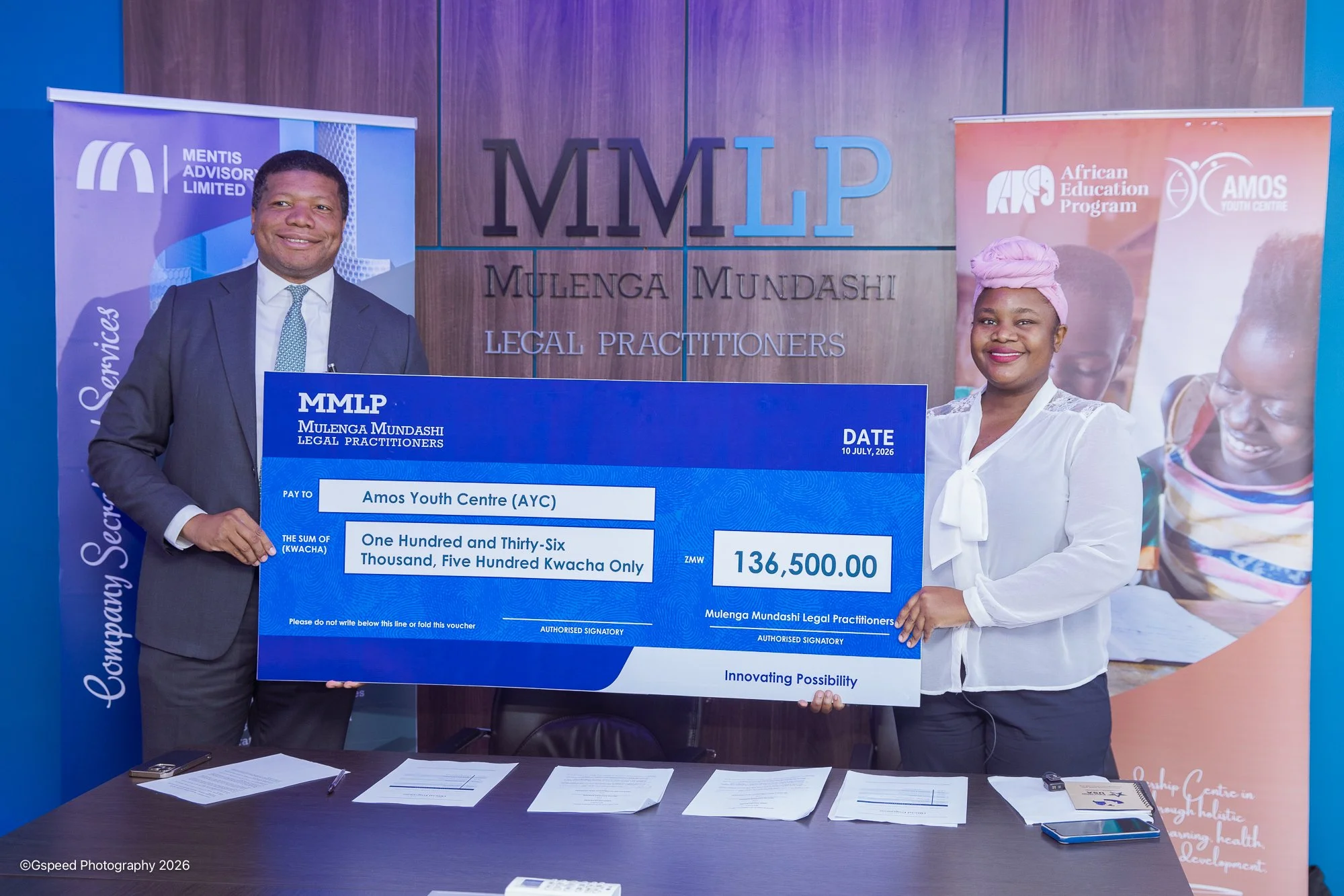 Two individuals, a man dressed formally in a suit and a woman in a white blouse, are holding a large ceremonial cheque together at an event. The man is smiling and the woman is also smiling. They are standing in front of a backdrop with logos and tex