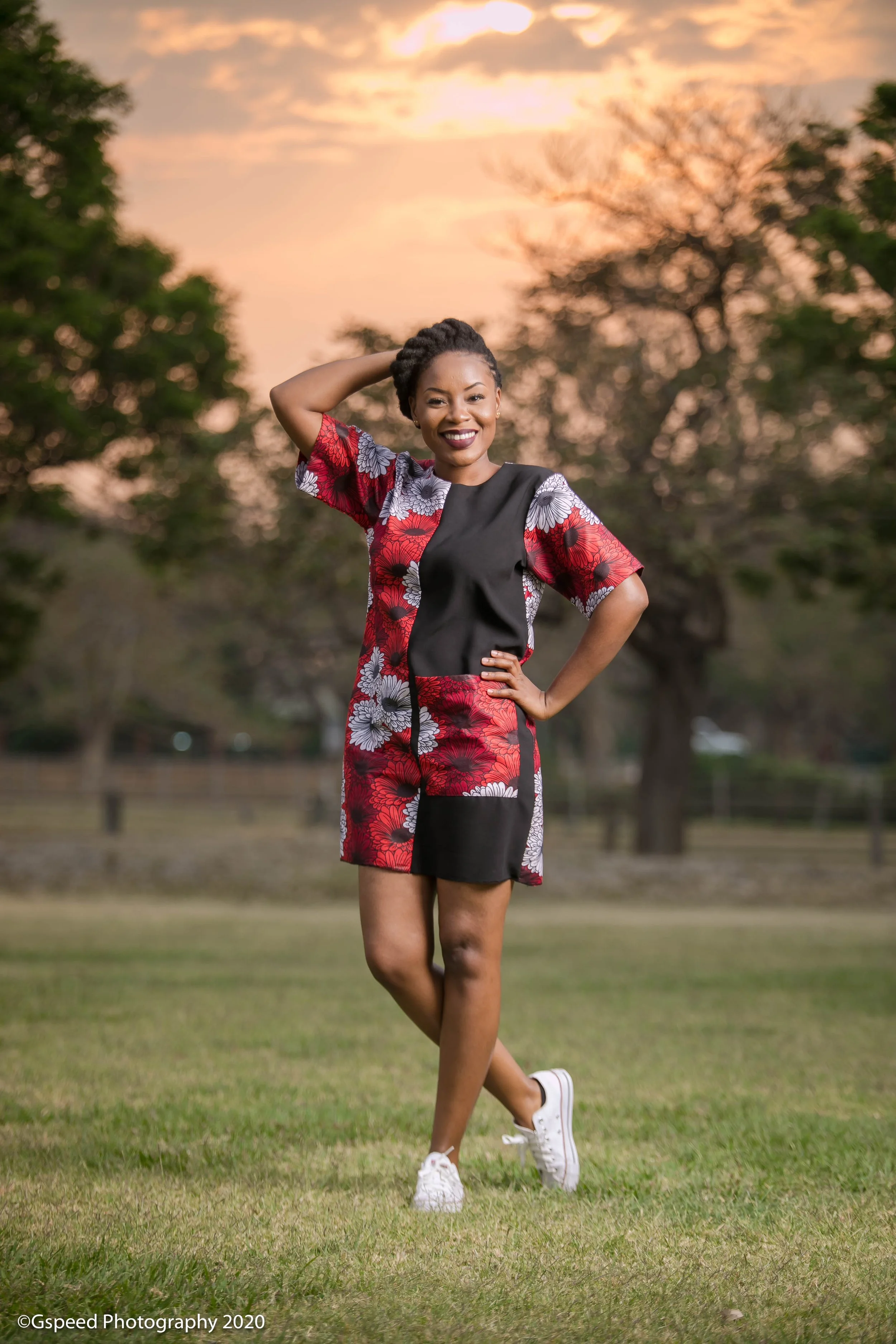 A woman with short, braided hair stands outdoors in a park during sunset, smiling and wearing a short dress with a floral pattern and white sneakers.
