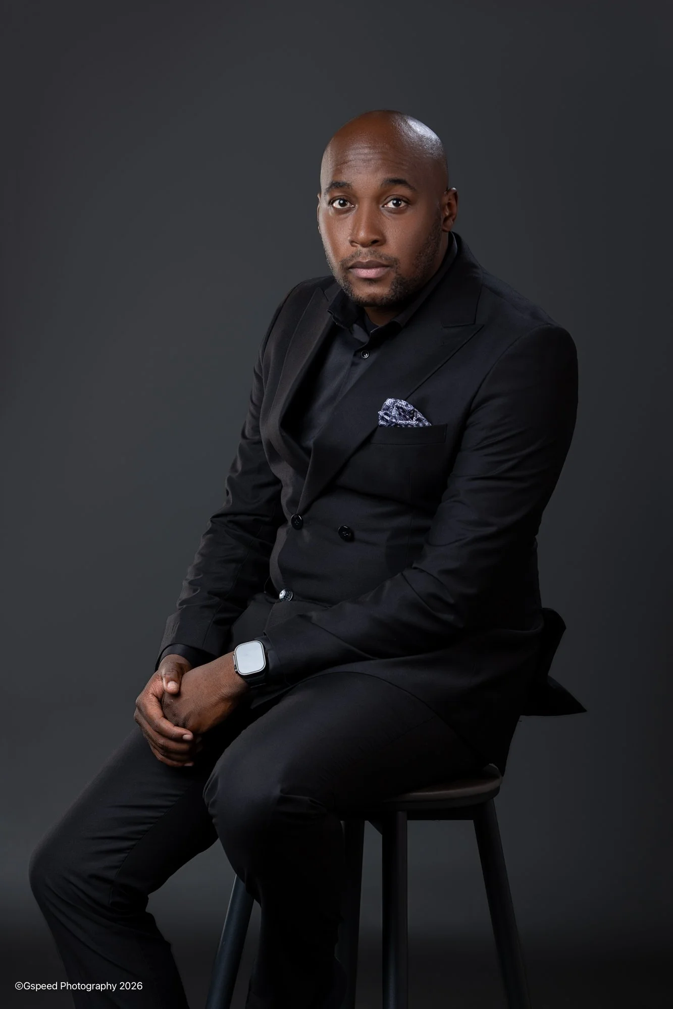A bald man in a black suit with a patterned pocket square sitting on a stool against a dark background.