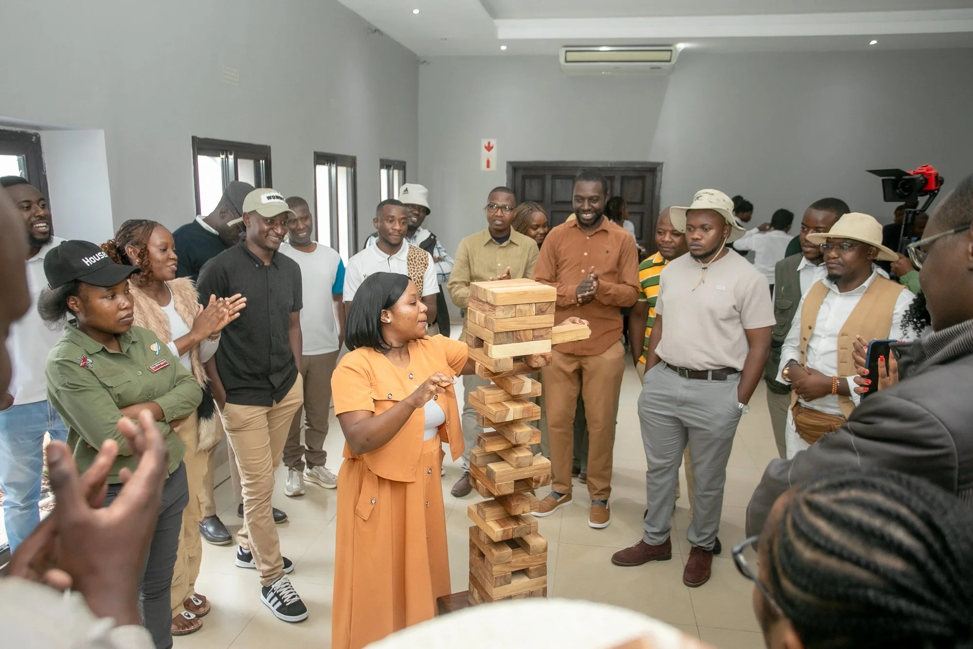A group of people gathered around a large Jenga game in a room with gray walls and multiple windows, some smiling and others watching as a woman in an orange dress is about to remove a wooden block from the Jenga tower.