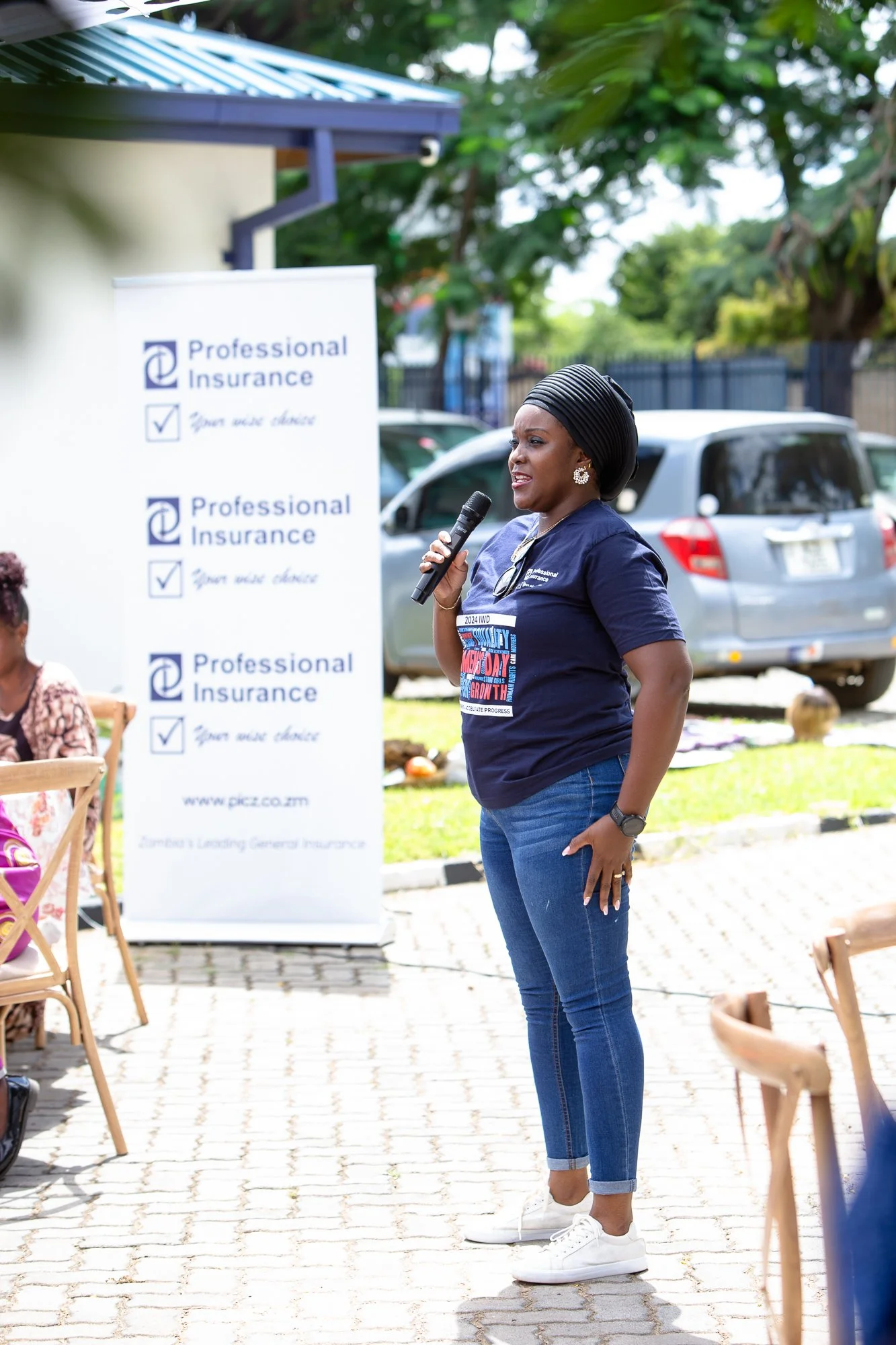 A woman wearing a navy blue t-shirt and jeans, holding a microphone, speaking outdoors at a professional insurance event, with a banner in the background.