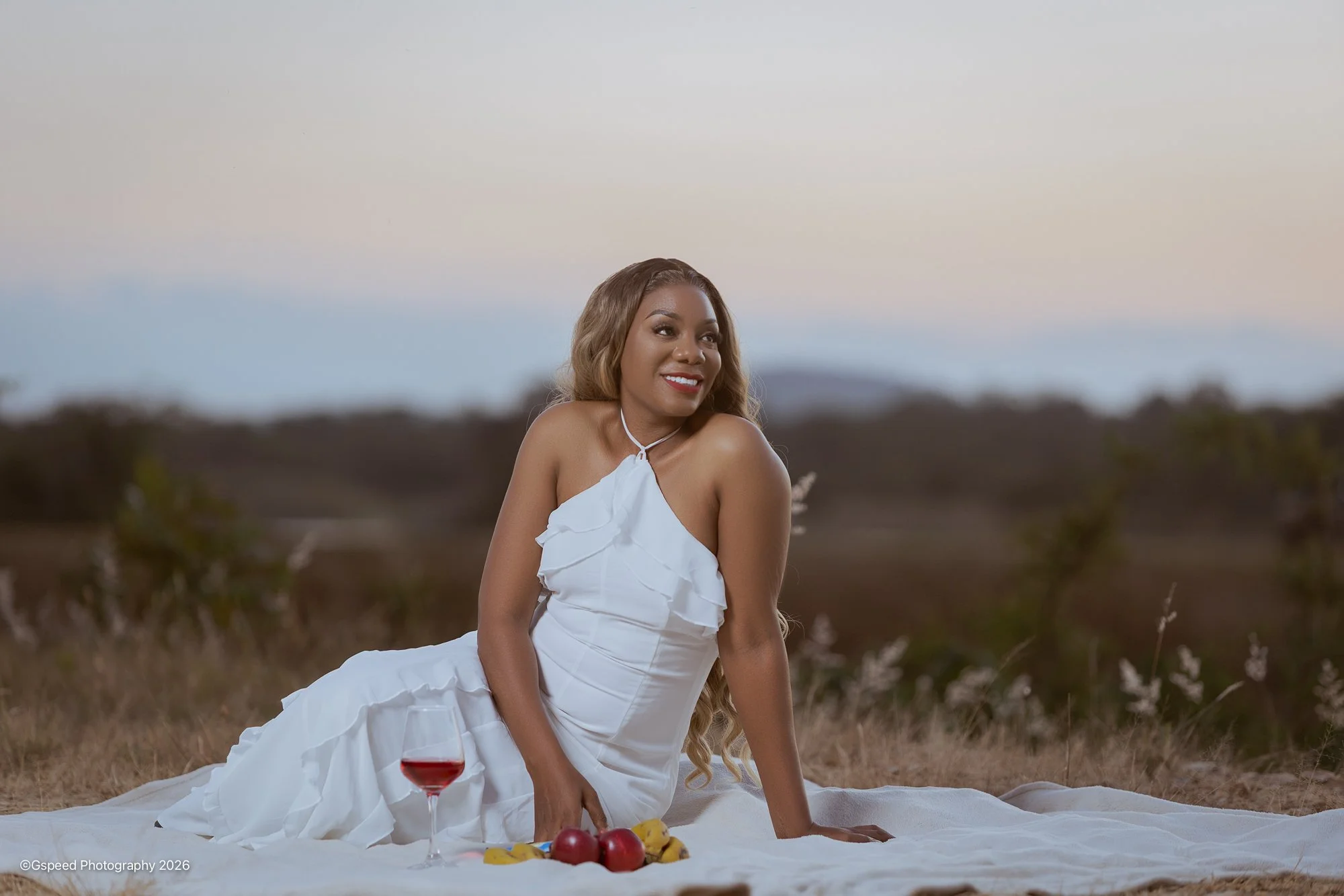 A woman with long blonde hair sits on a white blanket in an open field during sunset, wearing a white dress, smiling, with a glass of red wine and a plate of fruit nearby.