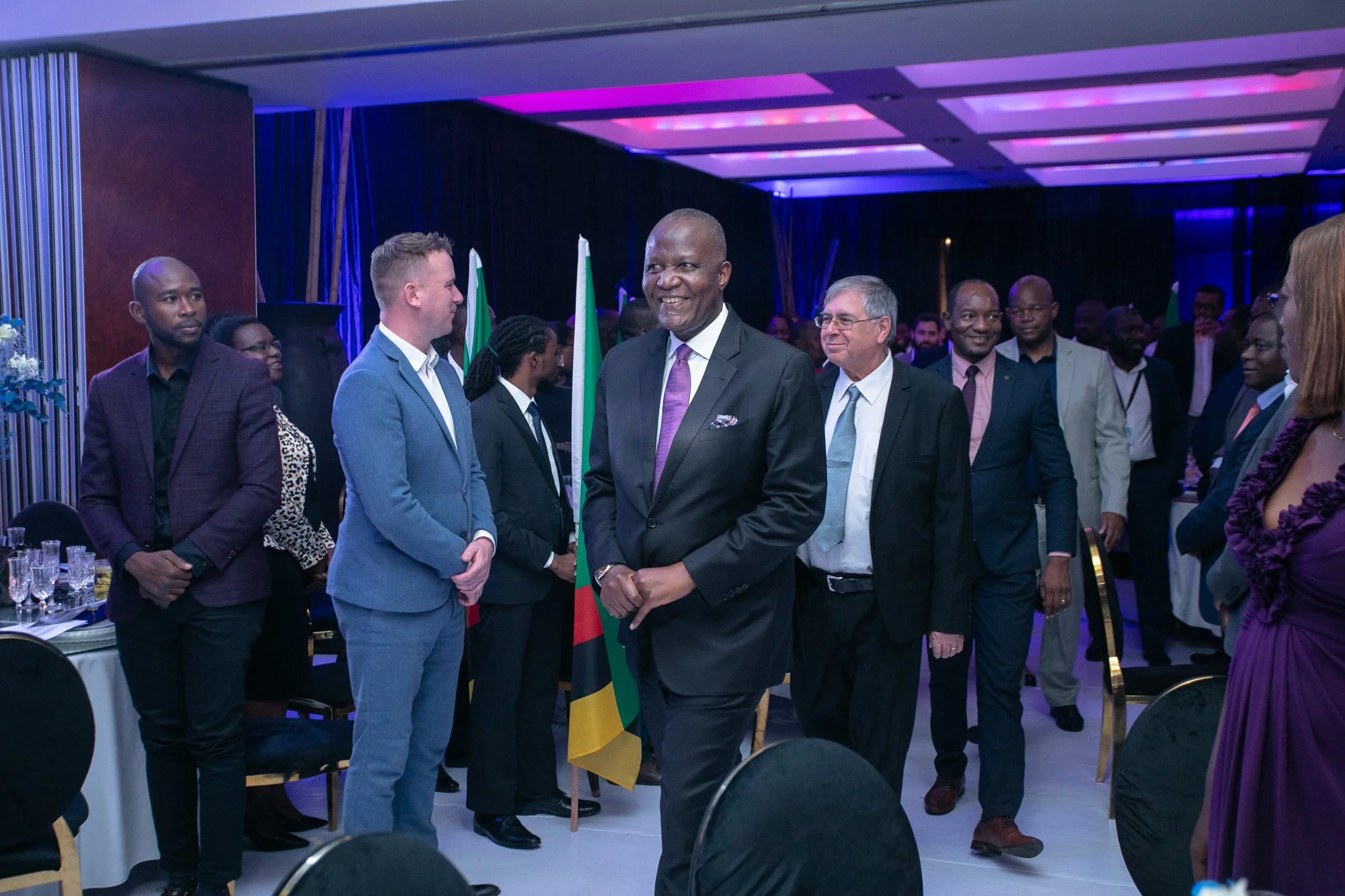 A group of business professionals standing and talking at a formal event with purple lighting, with South African flags displayed in the background. The central figure is a man in a dark suit, smiling and holding a South African flag.