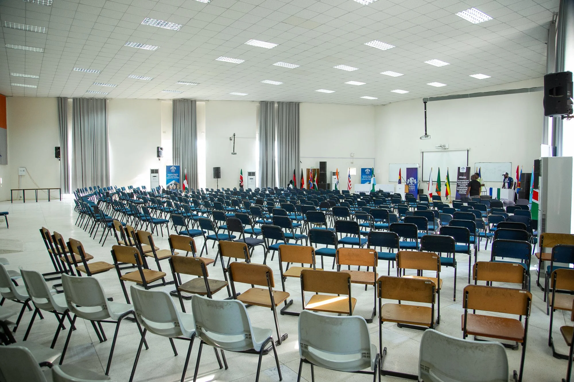 A large conference or seminar room set up with multiple rows of chairs facing a stage or presentation area. The room has high ceilings with numerous fluorescent lights, white walls, large windows with curtains, and audiovisual equipment including spe