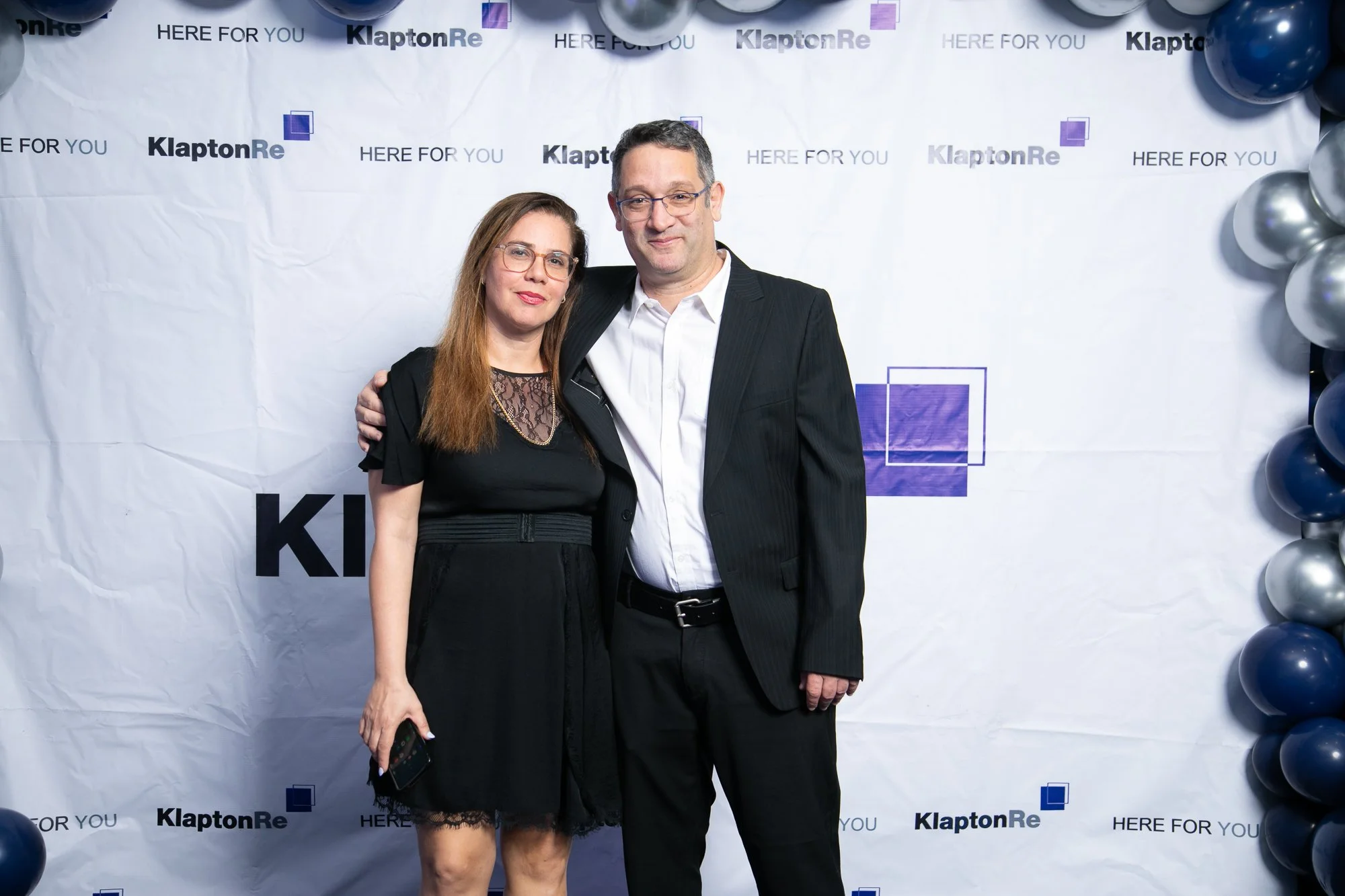 A man and a woman standing together at an event, both dressed in formal attire, with balloons and a backdrop that reads 'KlaptonRe' and 'HERE FOR YOU.'