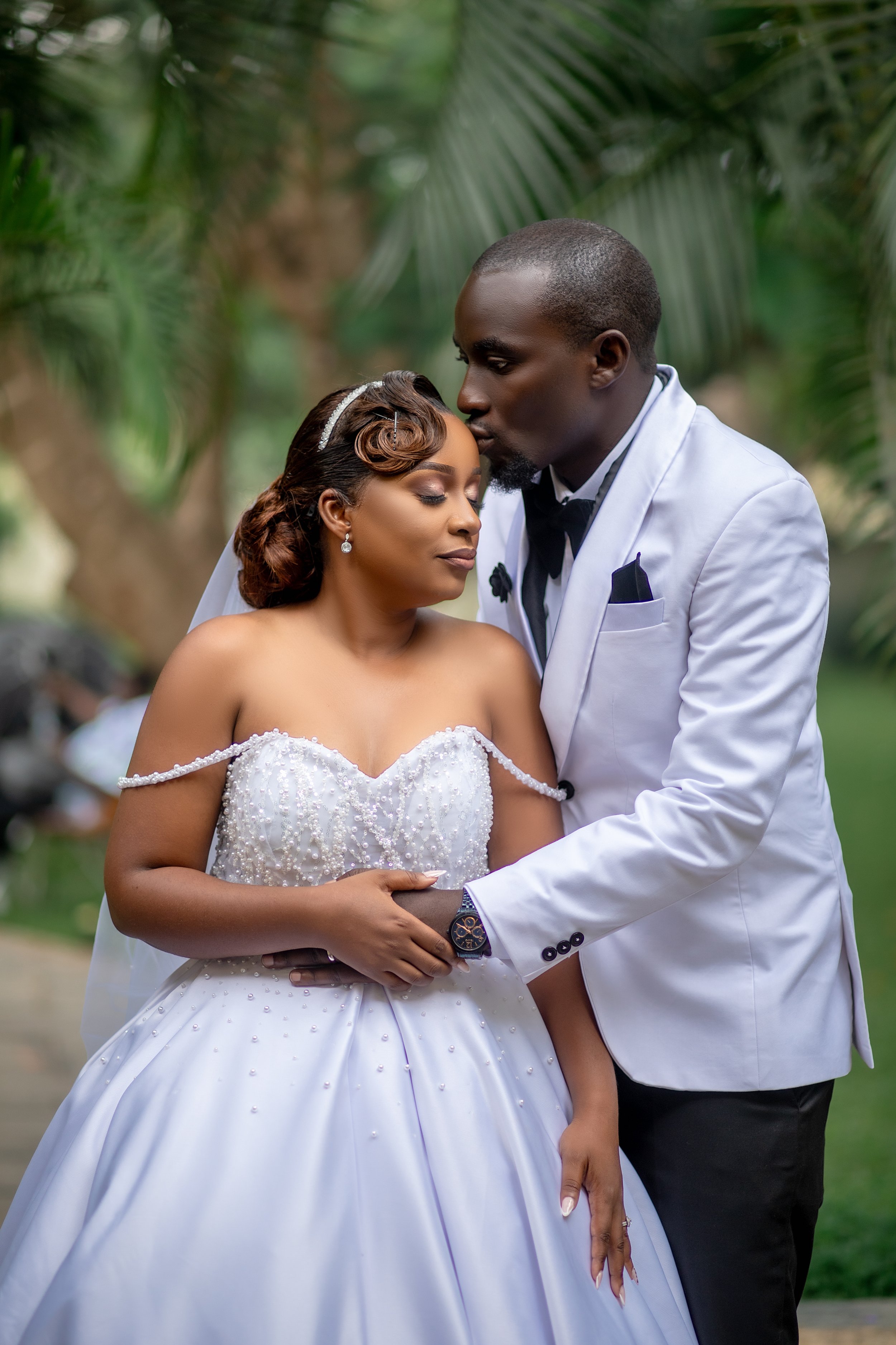 A bride and groom sharing a tender moment outdoors, with the groom kissing the bride's forehead. The bride is wearing a white wedding gown with pearl details and a veil, and has styled hair with a headband. The groom is dressed in a white tuxedo jack