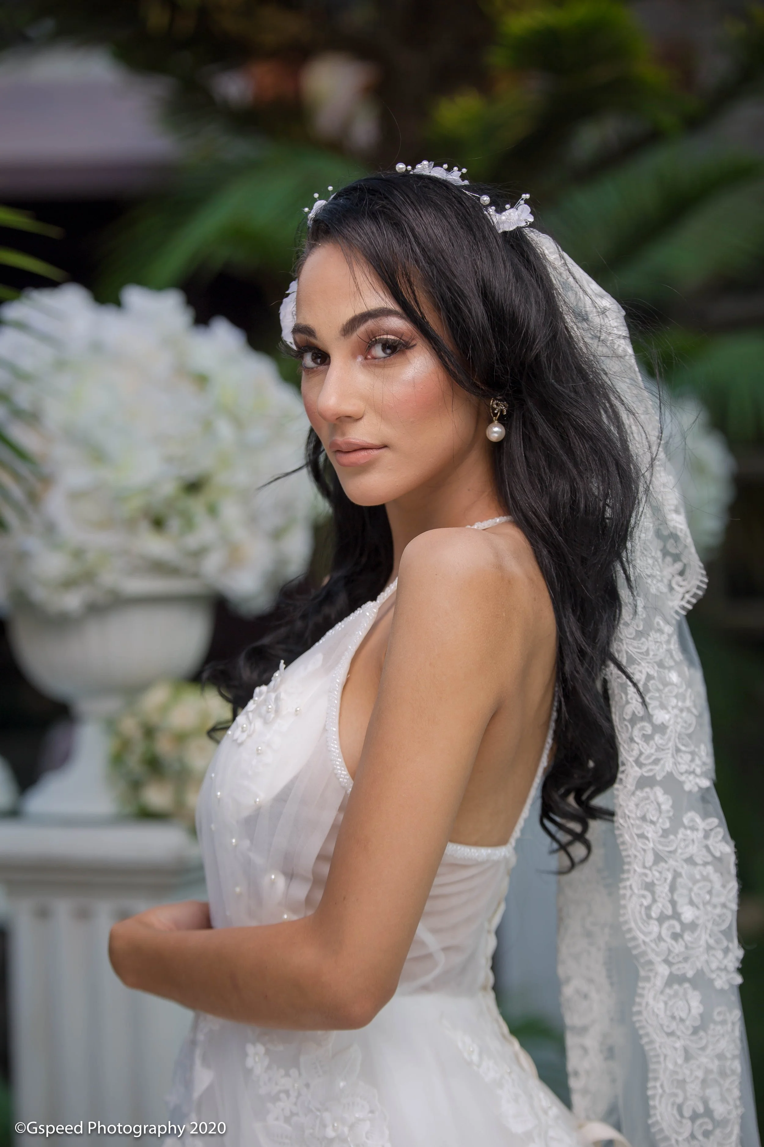 Close-up of a woman with dark hair wearing a wedding dress and pearl earrings, holding her hand, with a blurred background featuring white flowers and greenery.