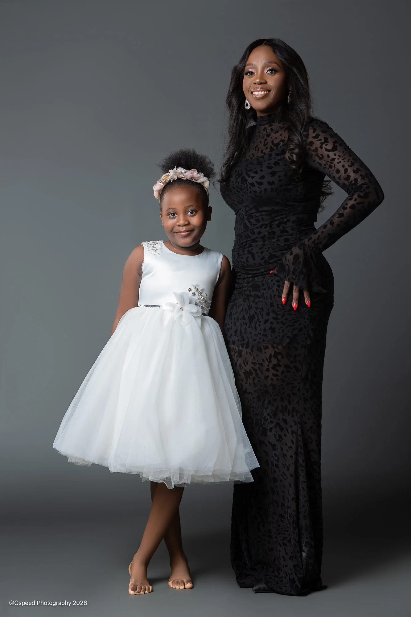 A woman and a young girl standing together against a plain grey background. The woman is wearing a long, black, leopard print dress with sheer long sleeves and red nail polish. The girl is in a white dress with a floral headband and is barefoot. Both