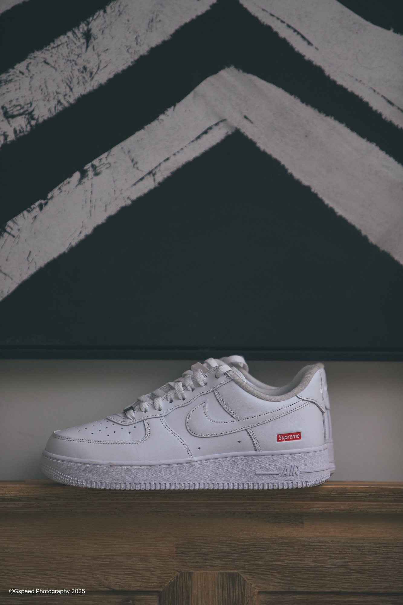 A white Nike Air Force 1 sneaker with a red Supreme logo on the side, placed on a wooden surface in front of a black box. The sneaker is viewed from the side, showing the Nike swoosh and details of the shoe.
