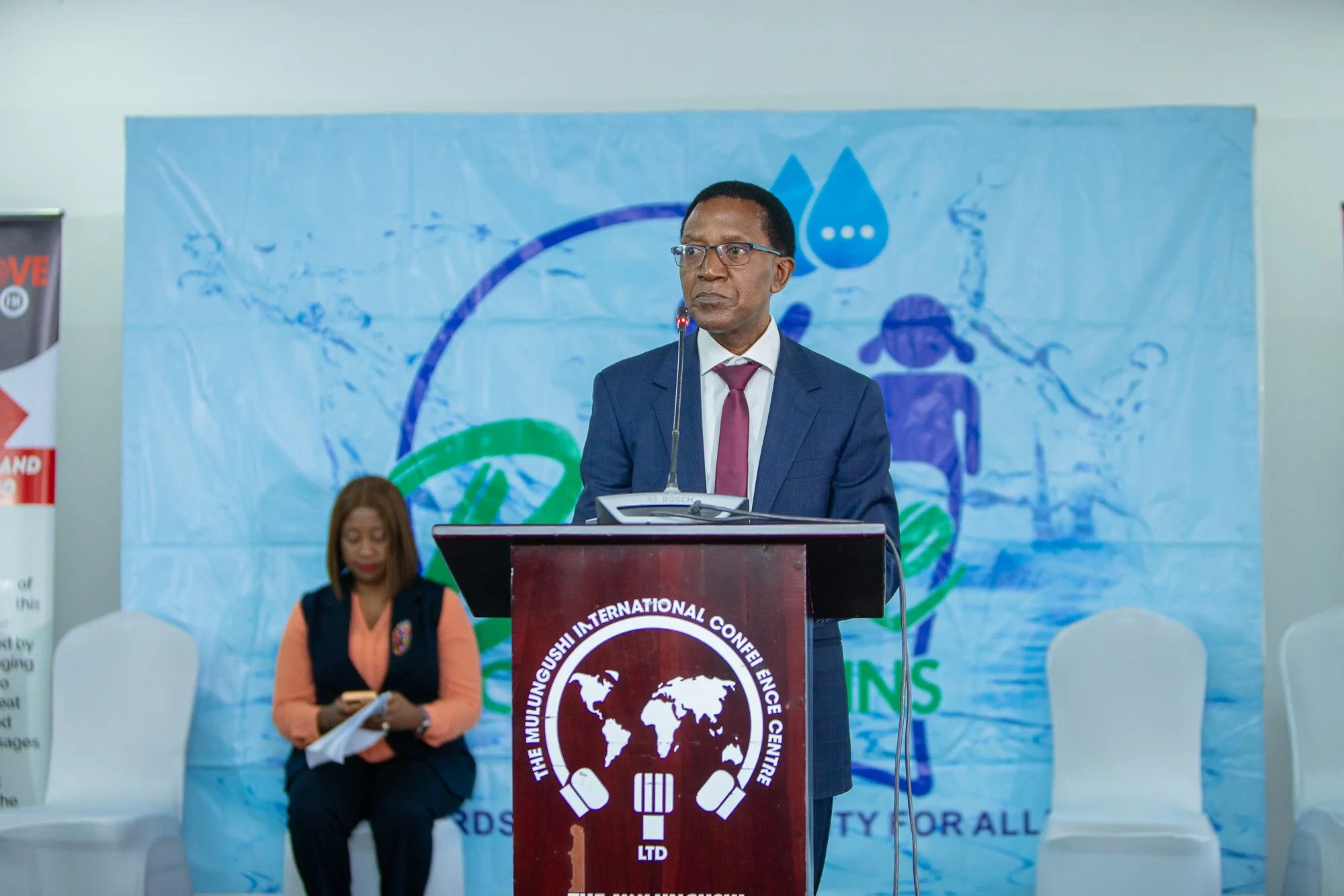A man in a suit and glasses stands behind a podium with a logo, speaking at a conference. A woman sits in front of him, holding papers and a phone, listening. There is a blue banner with a logo in the background.