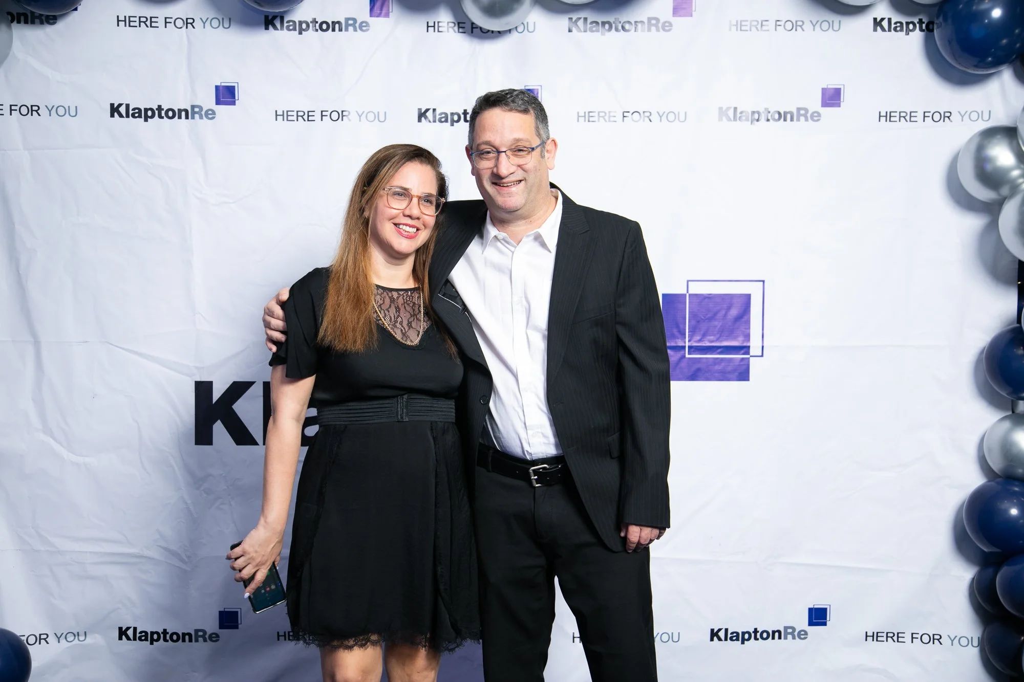 A man and a woman smiling and posing together at an event, with a background that features the words 'KlaptonRe' and 'Here For You' multiple times, decorated with balloons.