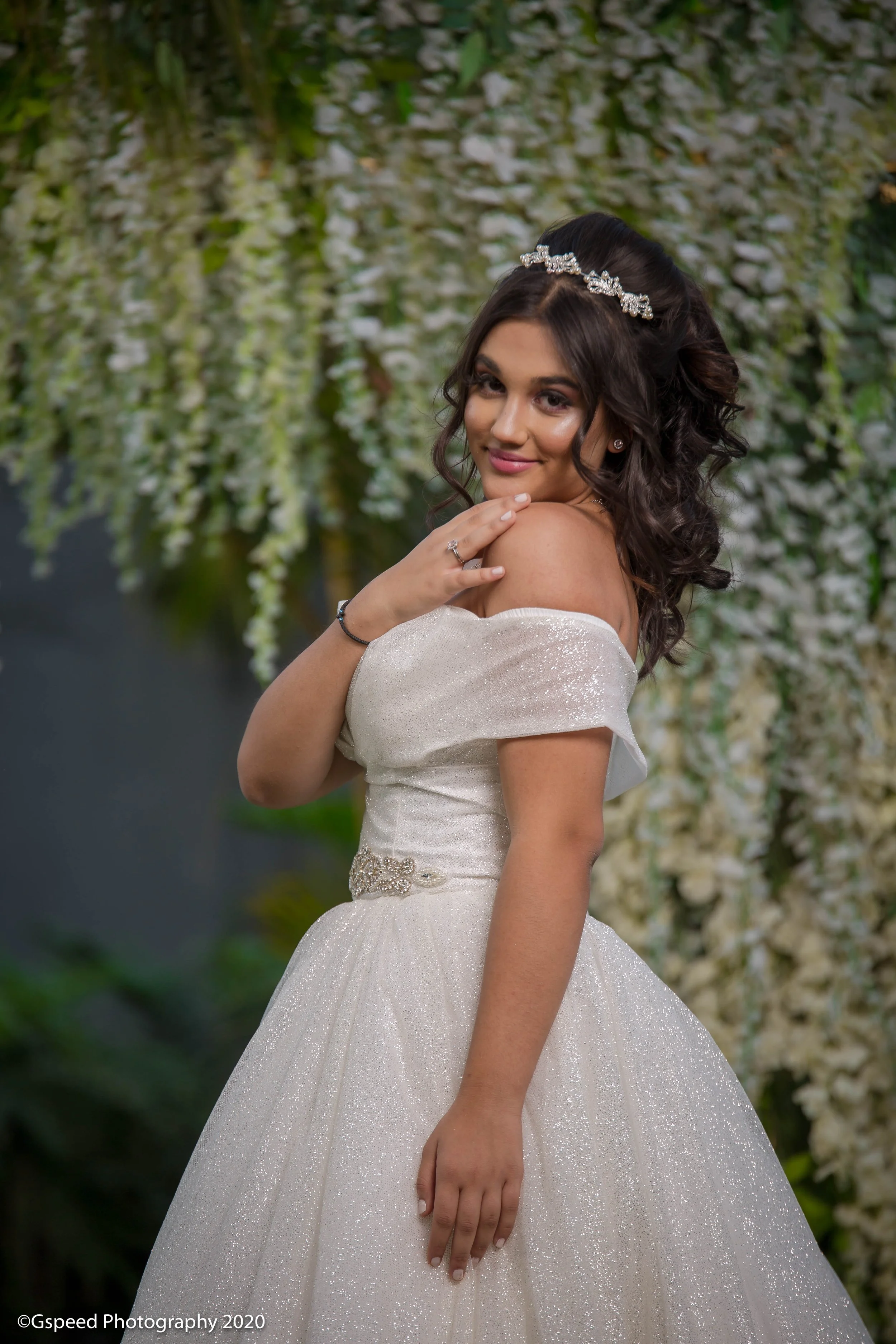 A young woman in a white, off-the-shoulder gown with a sparkling belt, wearing a silver tiara and jewelry, posing in front of a backdrop of cascading white flowers and greenery.