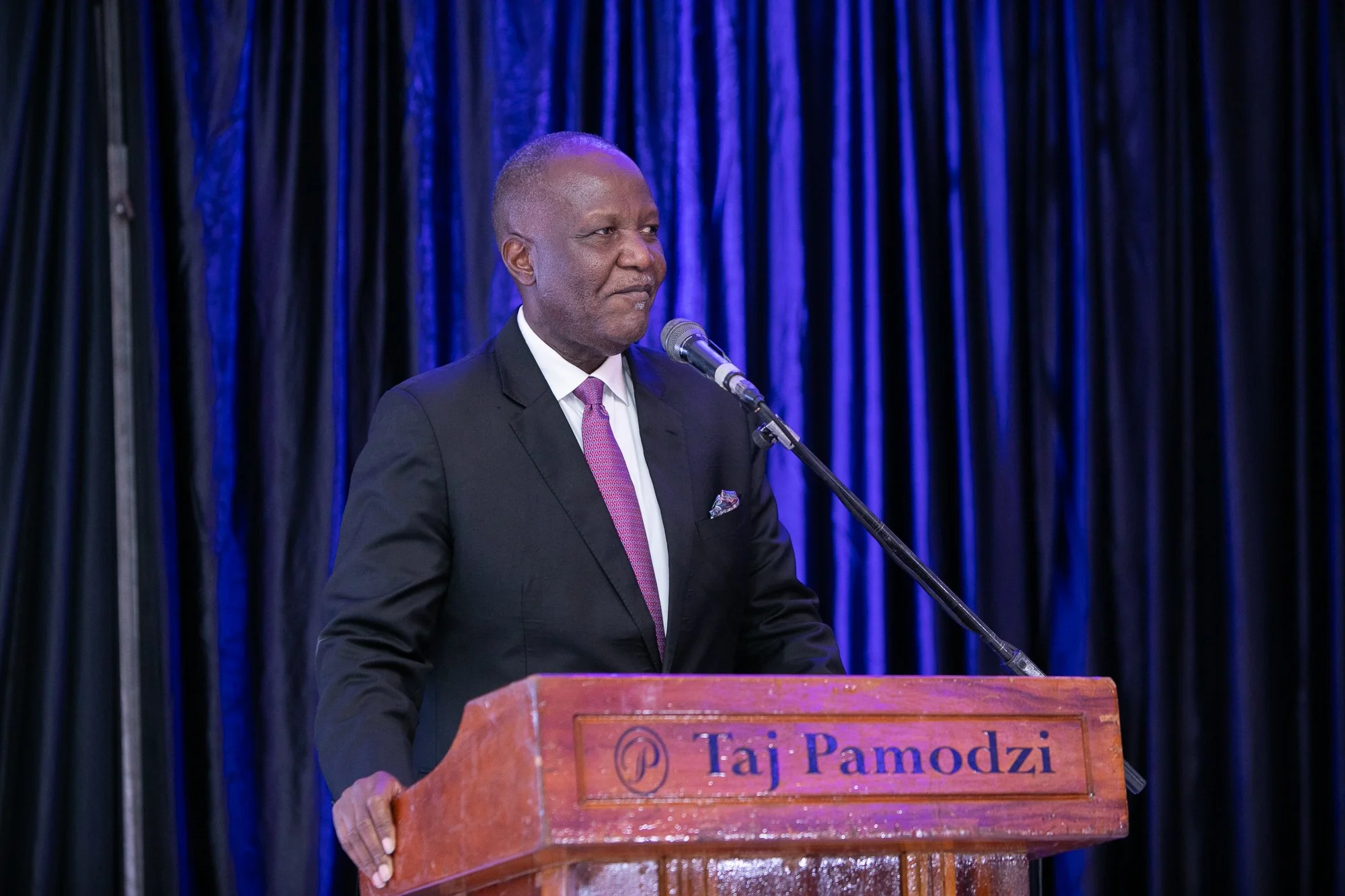 A man in a black suit and purple tie standing behind a podium with the name 'Taj Pamodzi' speaking into a microphone on stage with dark blue curtains in the background.