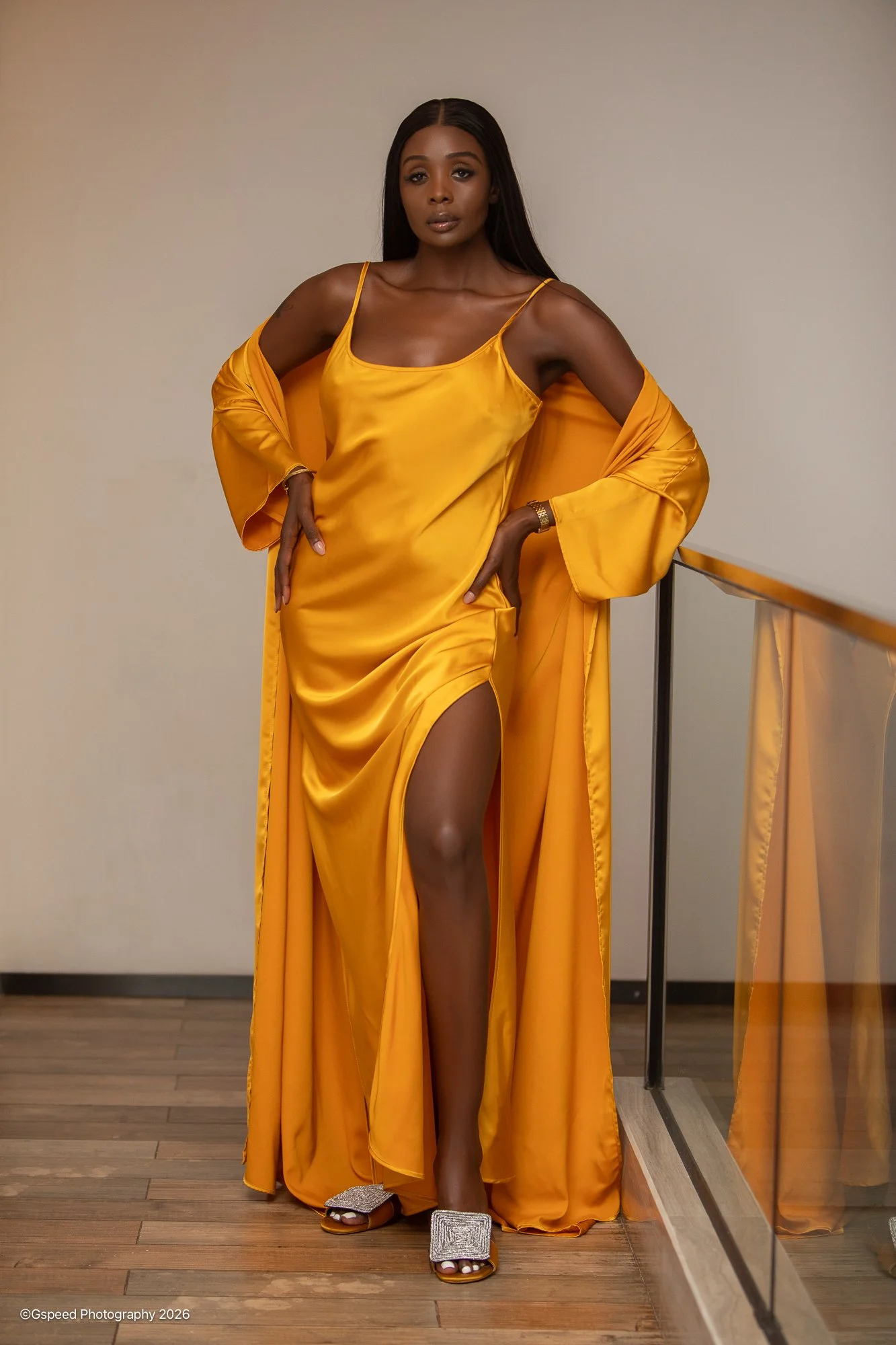 A woman with dark hair wearing a shiny yellow dress with a thigh-high slit, matching yellow robe, silver high-heeled sandals, and a watch, standing in front of a plain background.