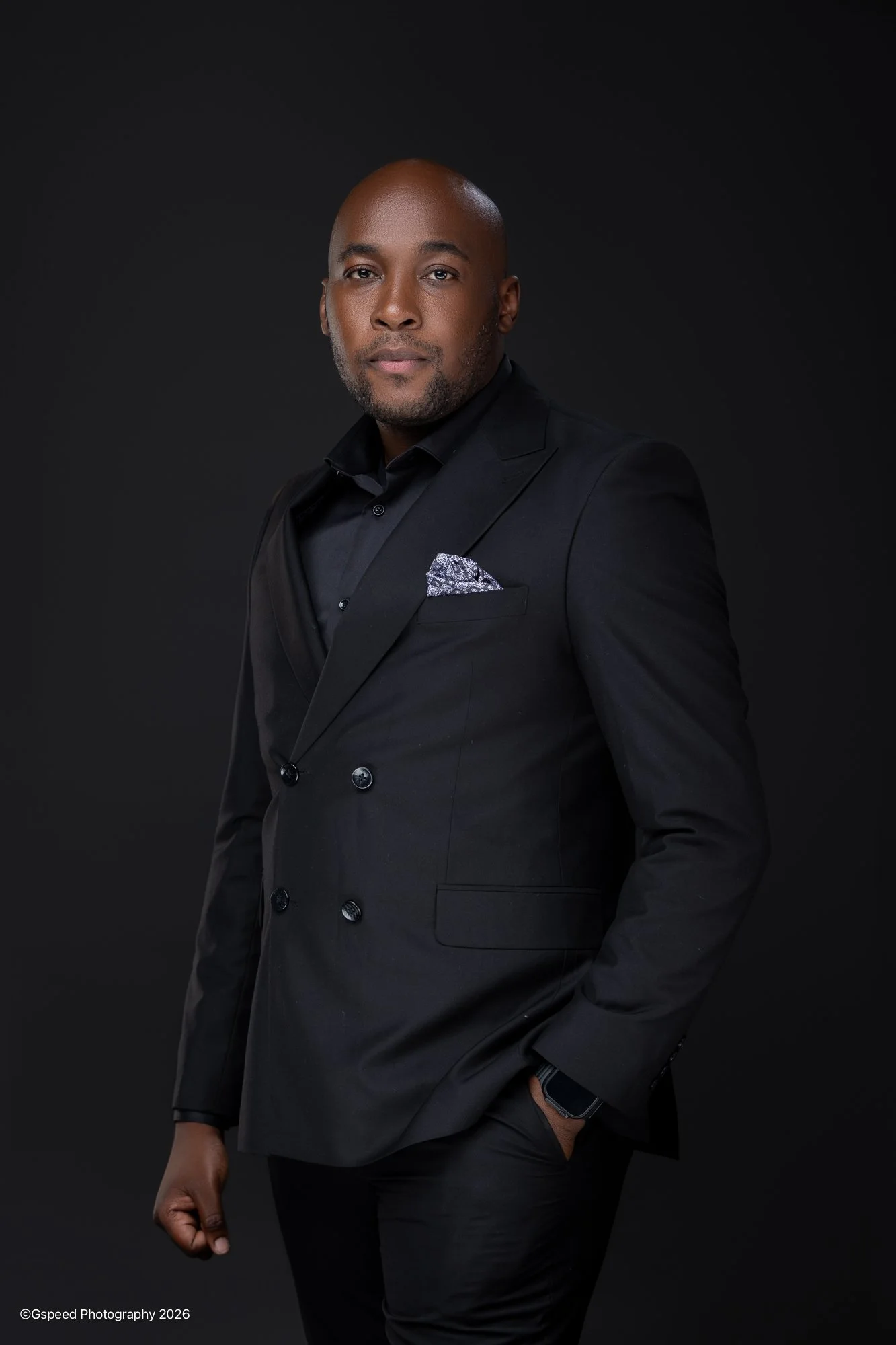 A man in a black double-breasted suit with a patterned pocket square, black shirt, and smartwatch posing against a dark background.