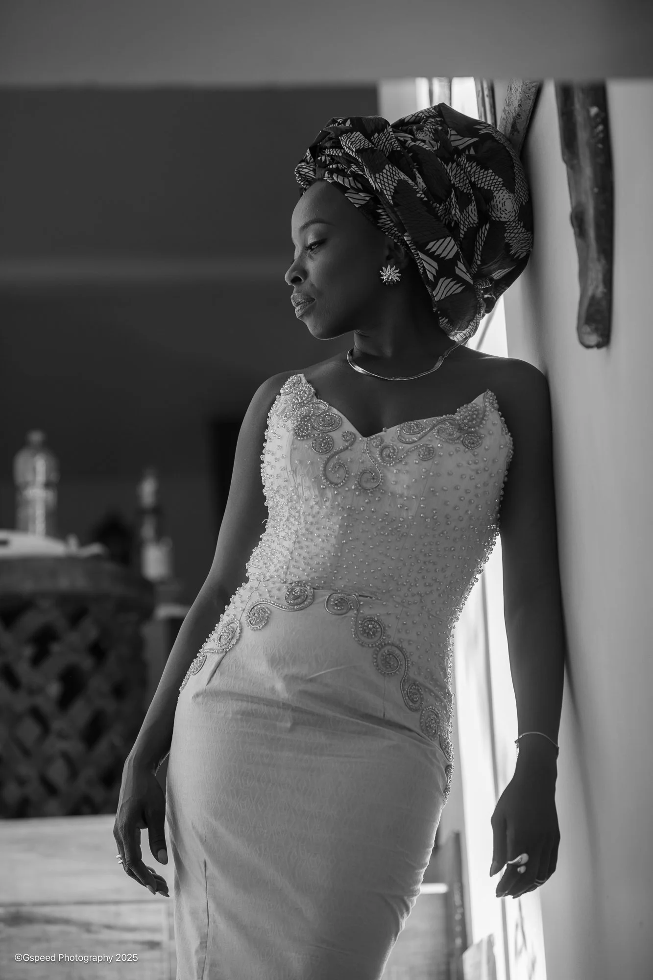A black and white photo of a woman wearing a detailed, embroidered dress and a patterned headwrap, standing against a wall with her eyes closed, looking serene.