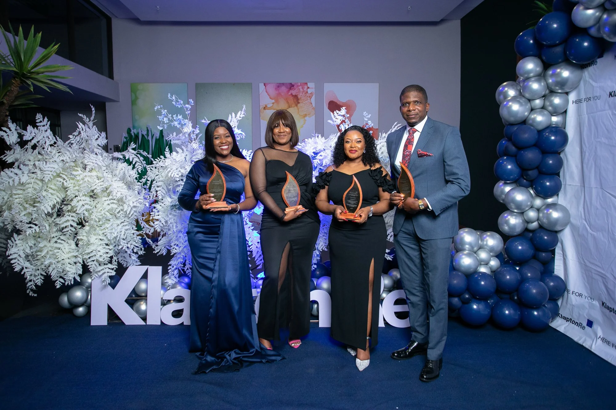 Four people in formal attire holding awards at an event decorated with white and blue balloons and artwork on the wall.