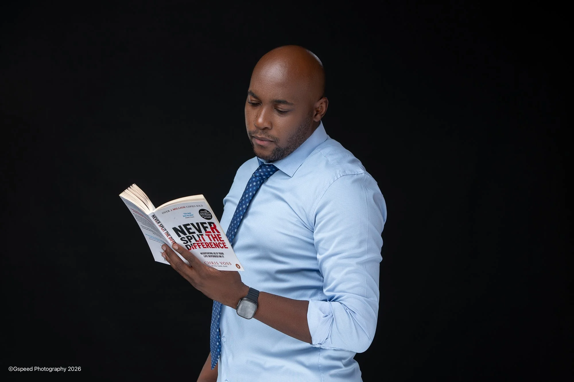 A man in a light blue dress shirt and patterned blue tie reading a book titled "Never Split the Difference" against a black background.
