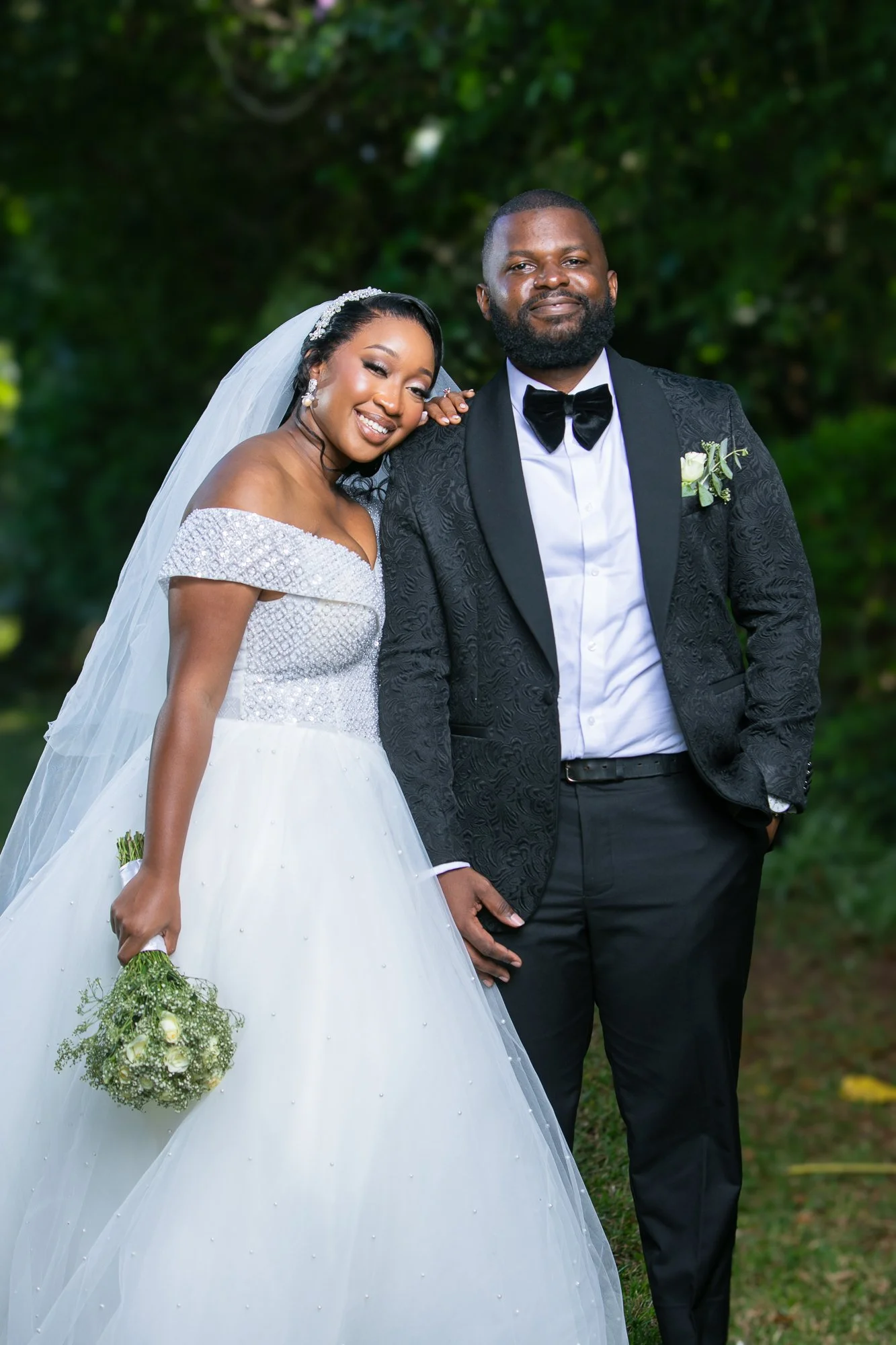 A bride and groom posing outdoors on their wedding day, with the bride smiling and holding a bouquet of flowers, and the groom standing confidently beside her with a serious expression. The bride is wearing a white off-the-shoulder wedding gown with 