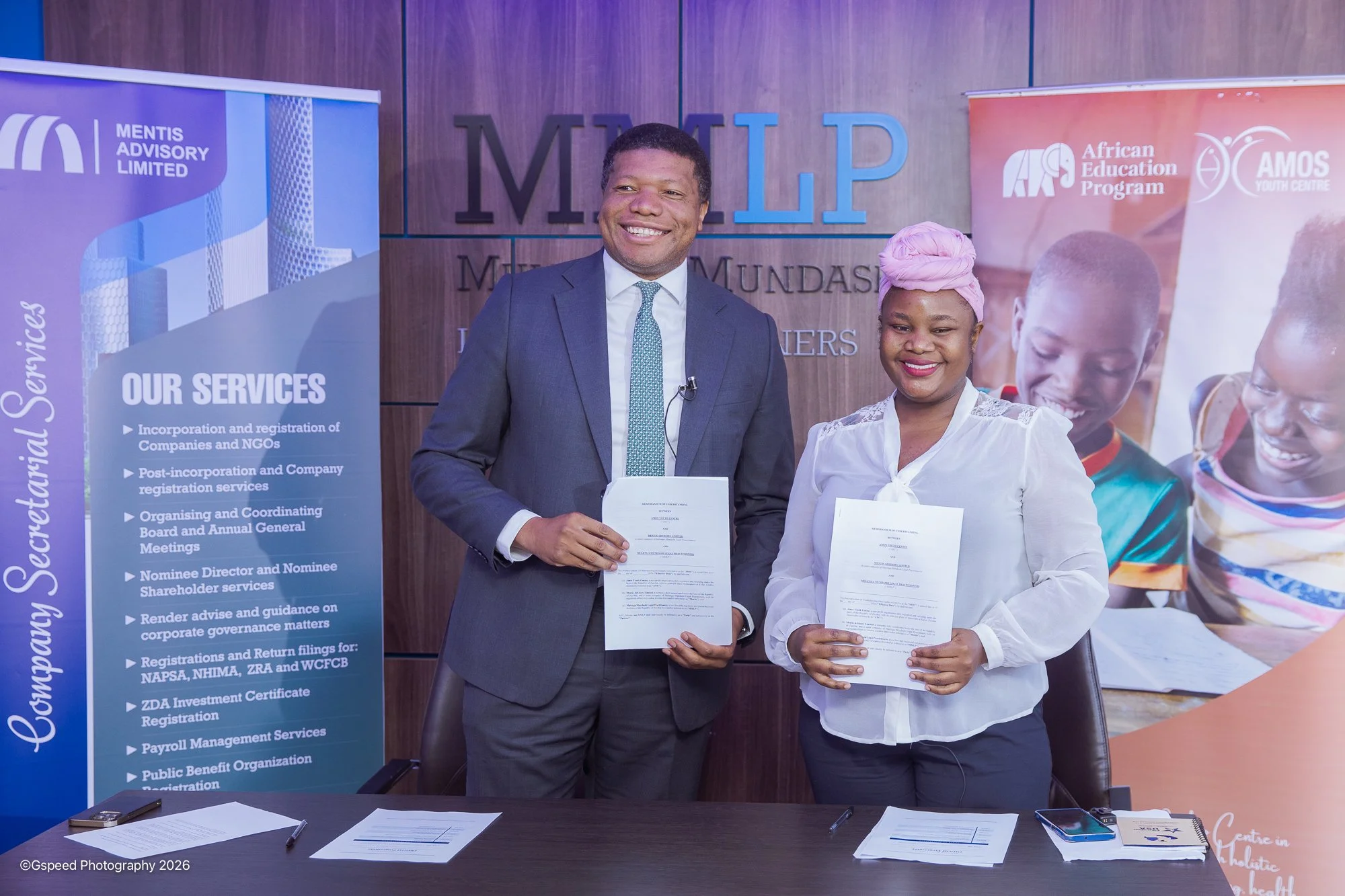 Two people, a man in a suit and a woman in a white blouse, standing indoors in front of banners and holding documents, smiling at the camera.