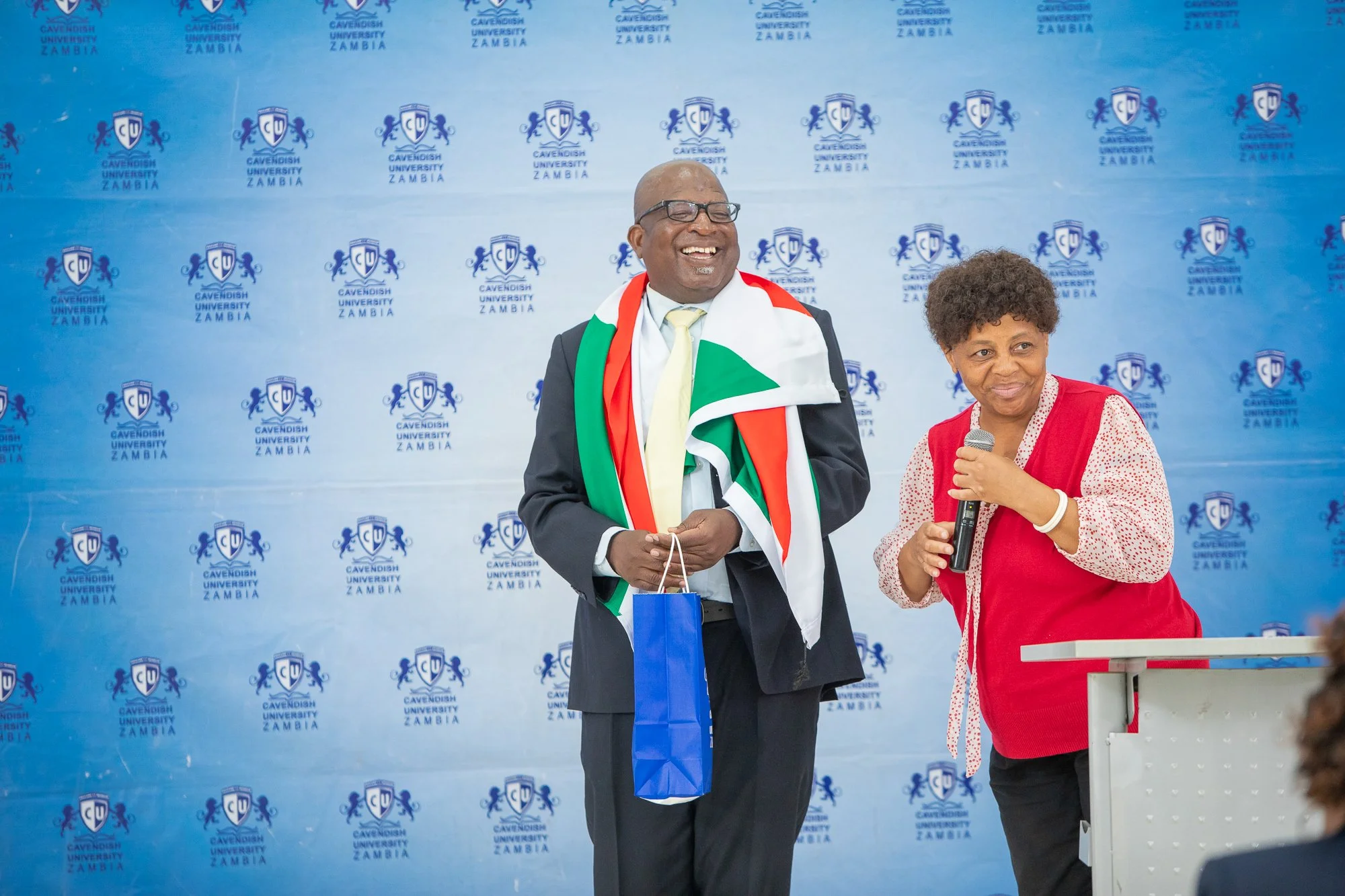 Two individuals standing in front of a backdrop with the logo of Cavendish University Zambia. The man is wearing a suit and glasses, with an academic gown with the Zambian flag colors draped over his shoulders. He is smiling and holding a blue gift b