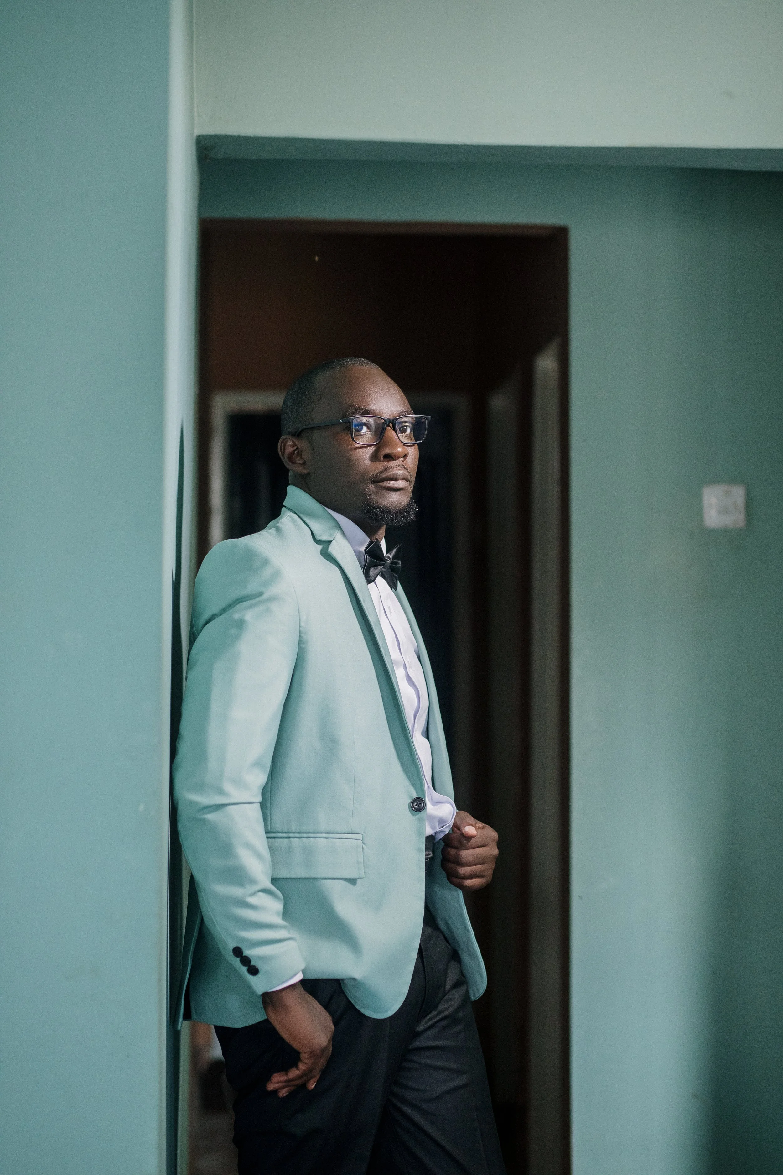A man wearing glasses, a light-colored suit, and a bow tie, standing in a doorway, looking to the side.