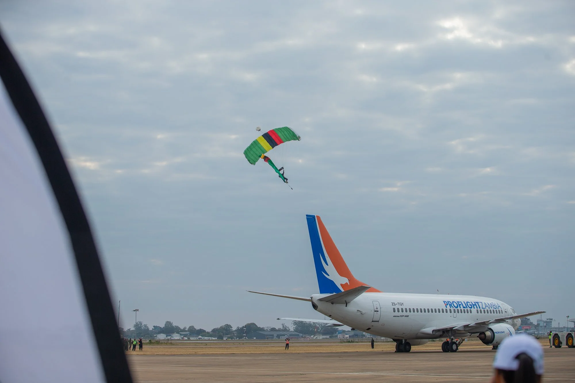 At an airport, a Zambia Airways airplane is parked on the runway, with a colorful parachute flying in the sky above and other aircrafts visible in the background.