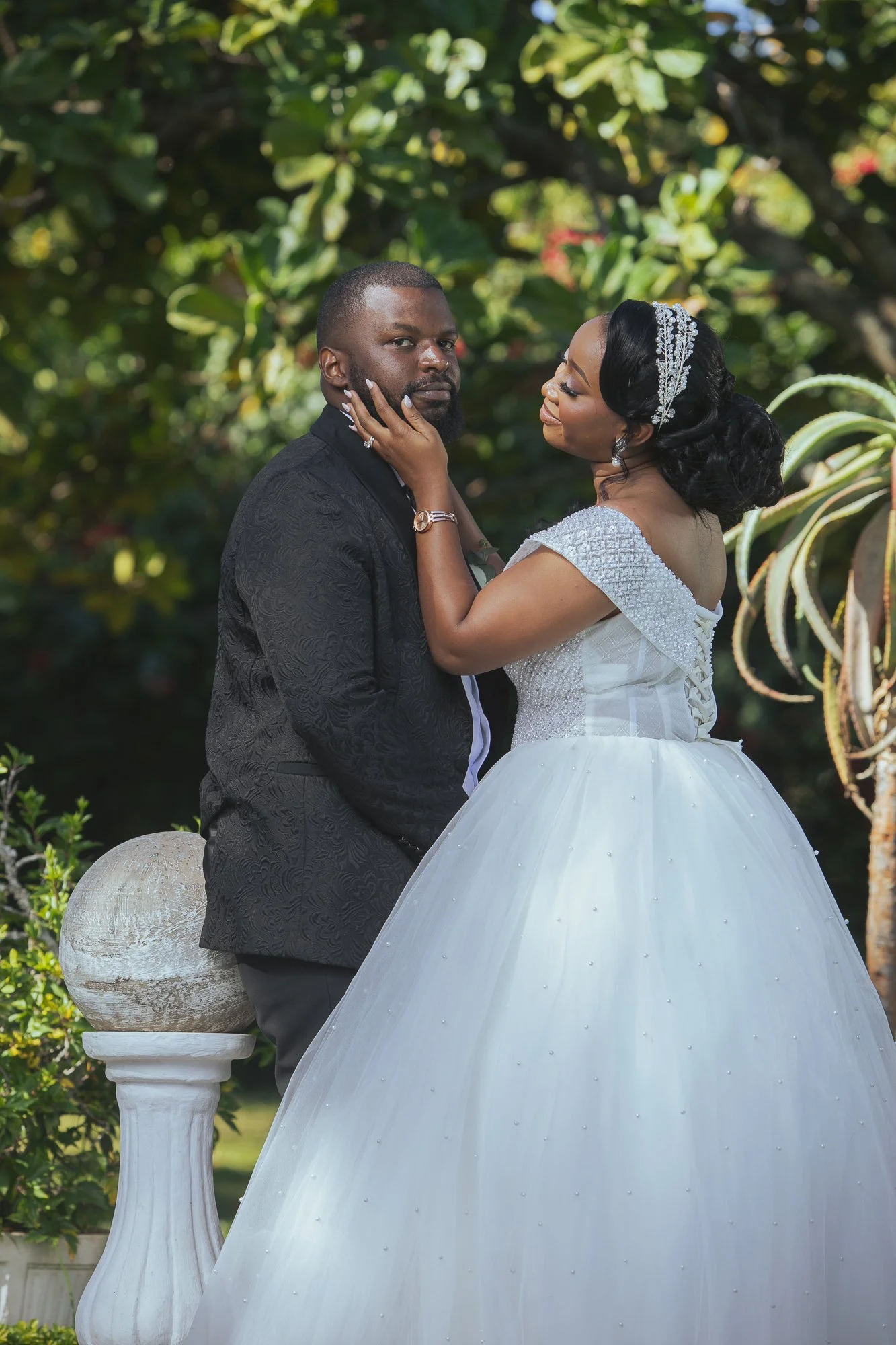 A bride and groom pose outdoors in front of lush green foliage. The bride is wearing a white gown with a beaded bodice and a tiara, her hair styled in elegant waves. The groom is dressed in a black suit with a patterned texture. The bride gently touc
