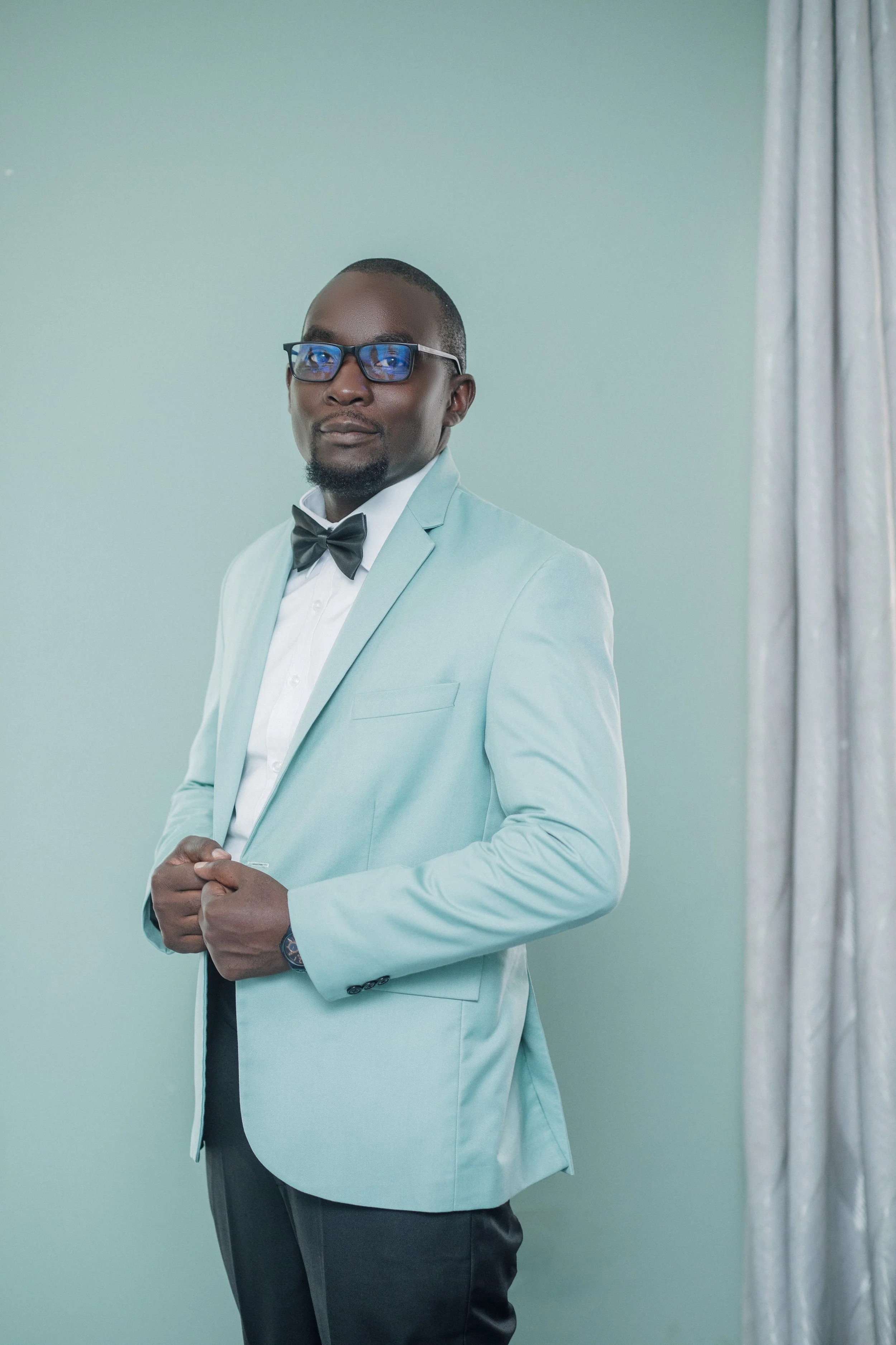A man dressed in a light blue tuxedo with a black bow tie and glasses, standing against a light green background with curtains on the side.