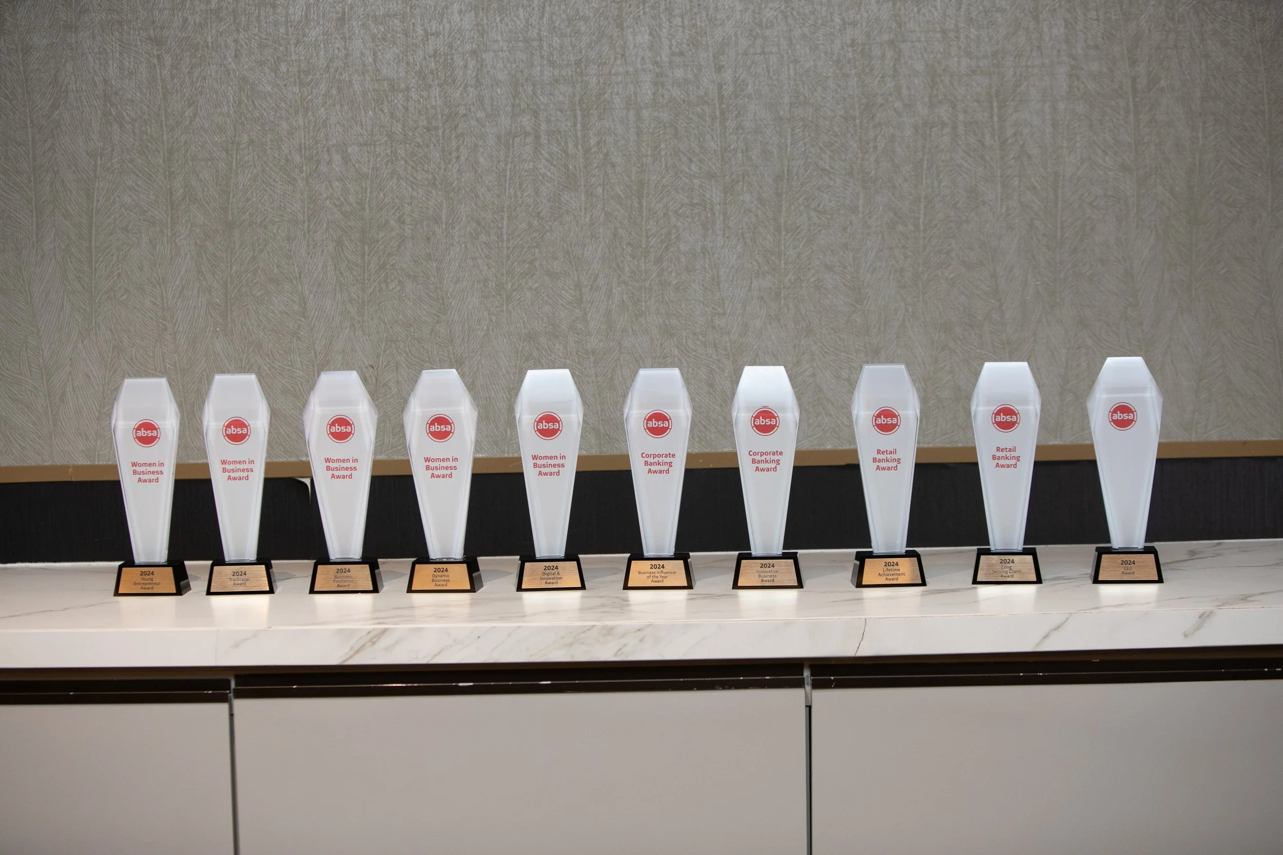 A row of acrylic awards with red and white logos and labels on a white marble surface, against a beige textured wall.