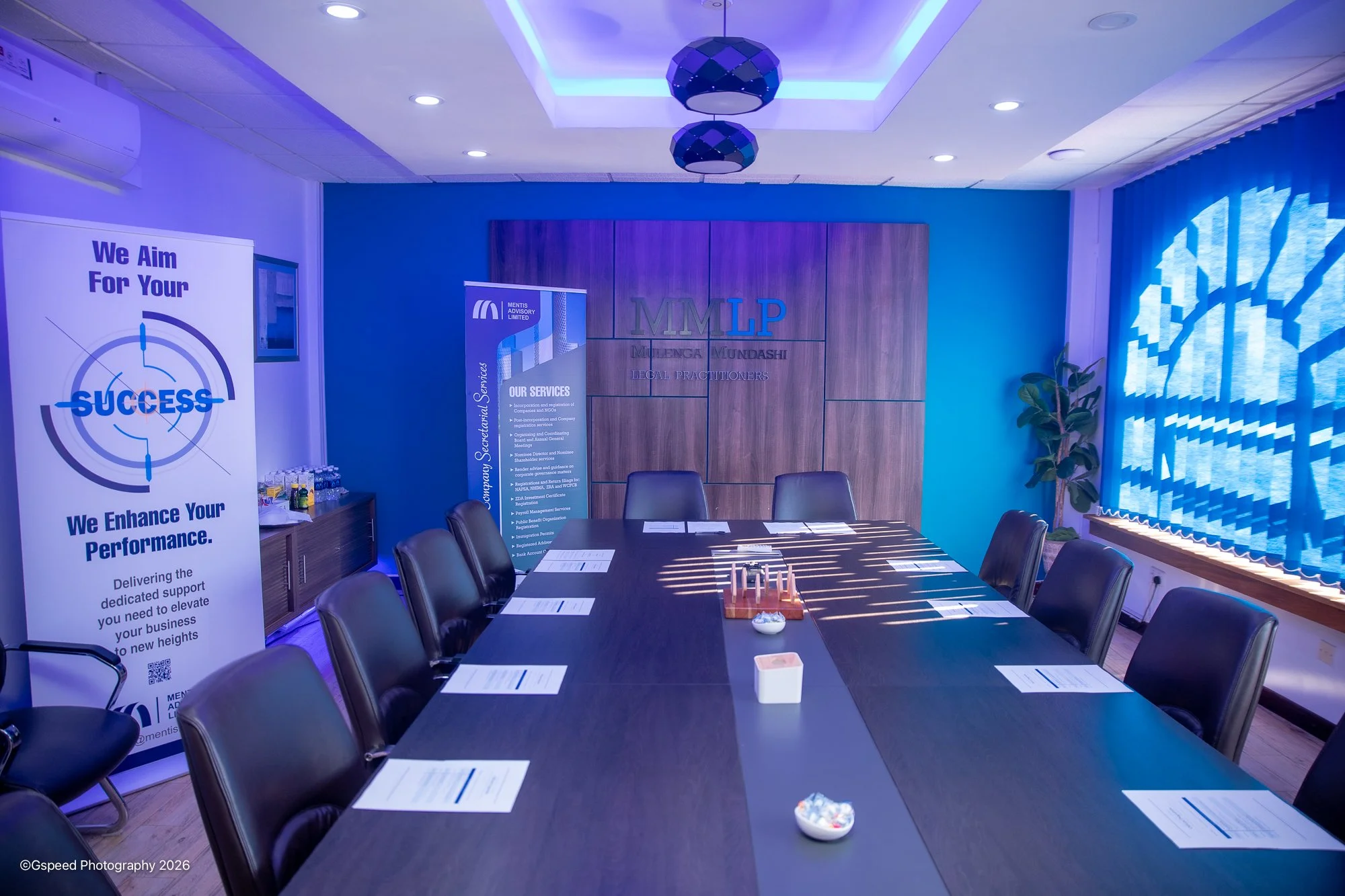 A conference room with a large dark table, surrounded by black chairs, papers, and small bowls on the table, with a window with blue curtains and a plant to the right, and promotional banners on the back wall for Mentis Advisory Limited with a wooden