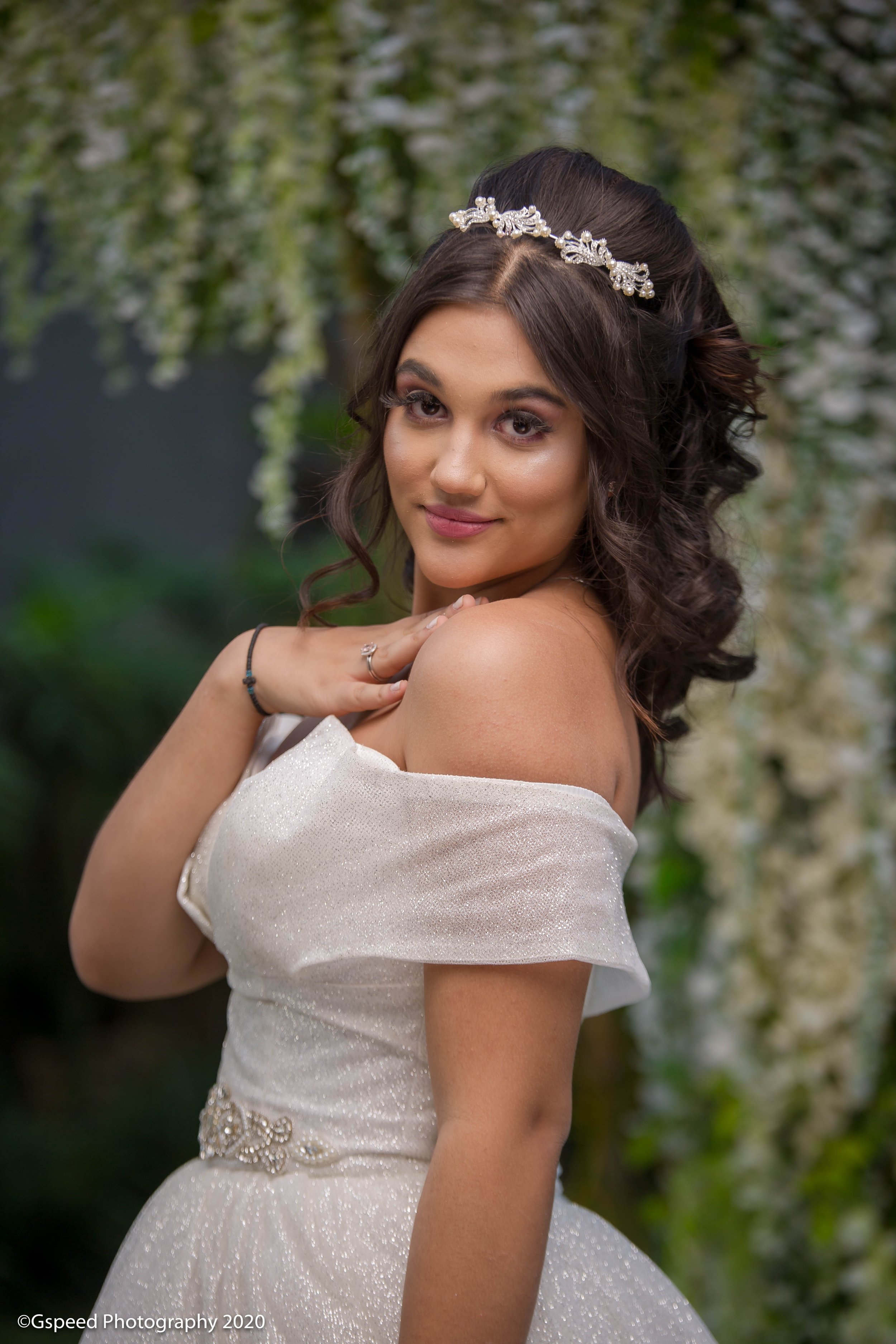 Portrait of a young woman with dark, styled hair wearing an off-the-shoulder white dress with a glittery fabric, a silver floral belt, and a decorative hairpiece, posing outdoors with greenery and white flowers in the background.