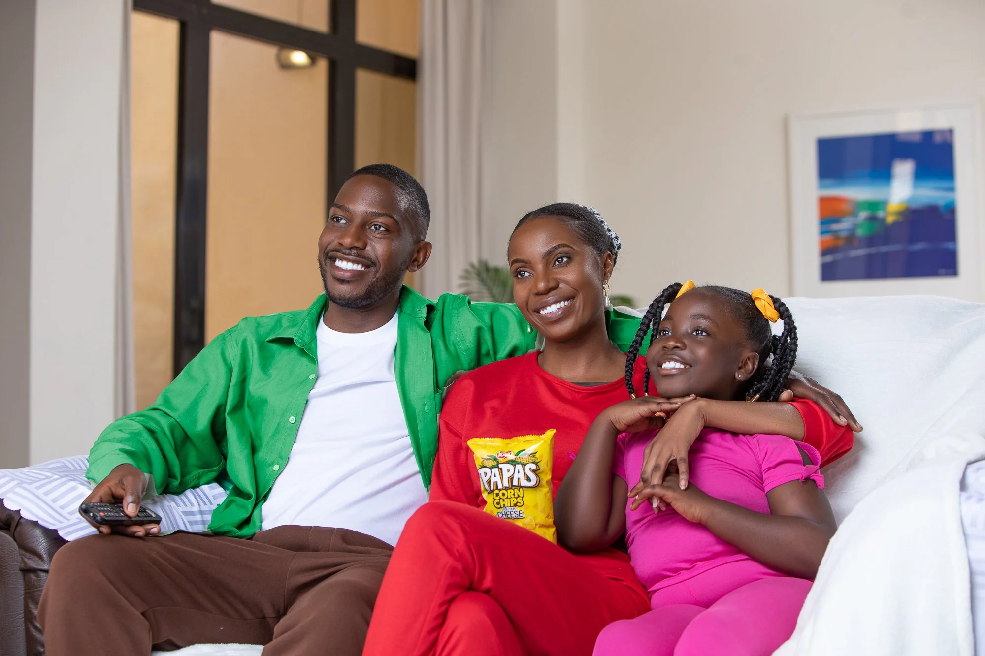 A family of three sitting on a couch watching TV happily, with snacks visible. The family includes an adult man, an adult woman, and a young girl, all smiling.