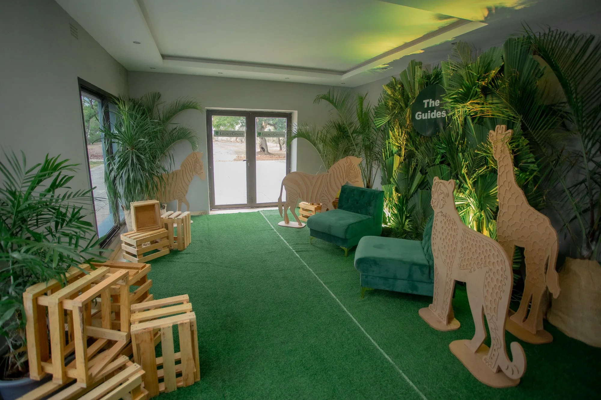 Indoor space with green artificial grass, potted plants, cardboard animal cutouts, and wooden crates, with glass door and window leading outdoors.