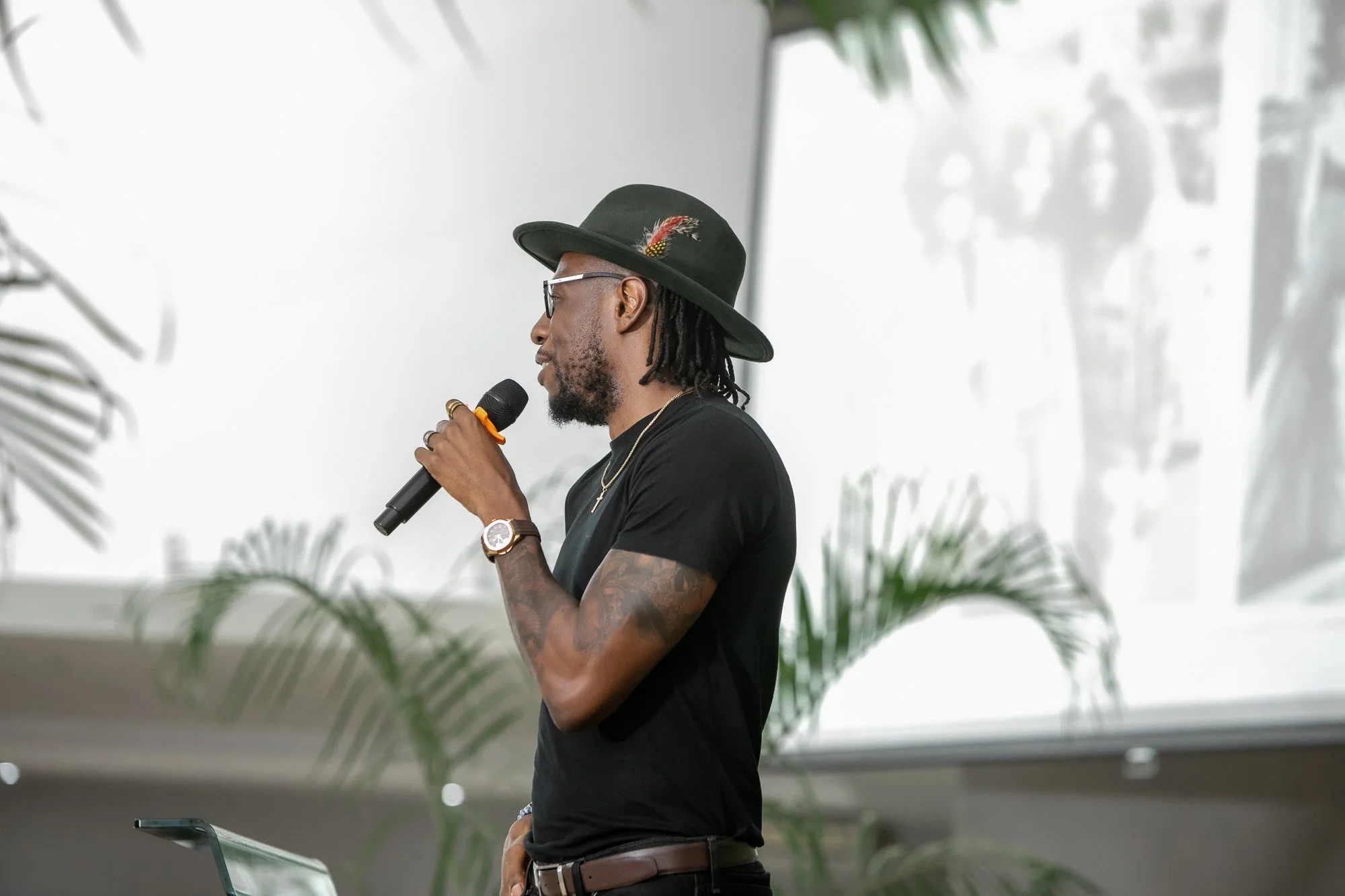 A man with tattoos wearing a black hat with a feather, glasses, and a black t-shirt speaking into a microphone during an event with plants and a white wall in the background.