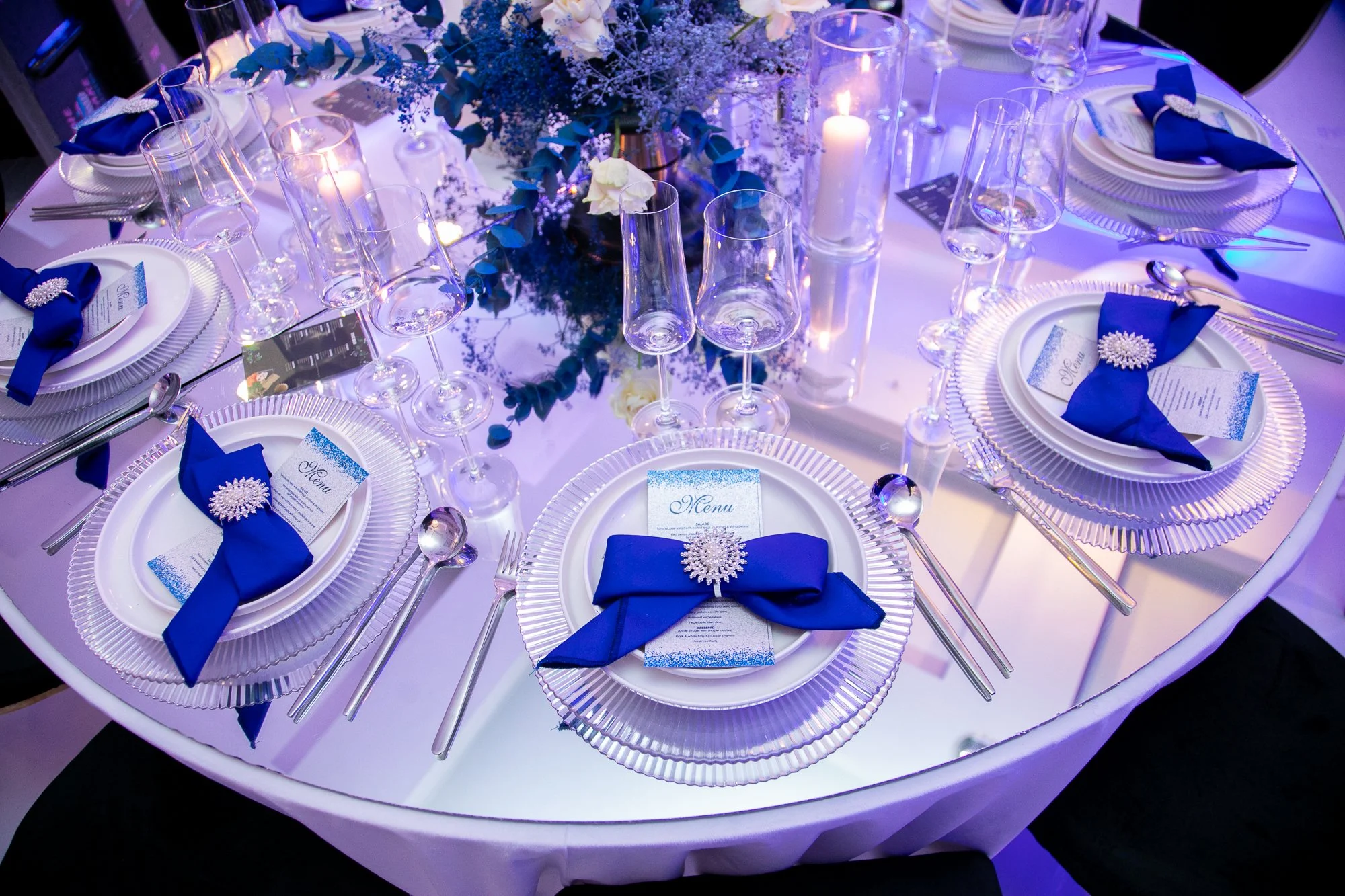 Round table decorated for a formal event with white tablecloth, elegant glassware, and blue napkins folded with decorative brooches. The centerpiece features purple and white flowers with candles in glass holders, creating a sophisticated ambiance.