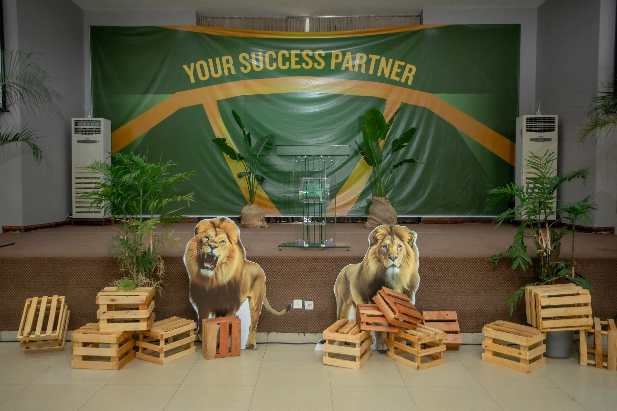 Stage setup with a green backdrop displaying the phrase 'Your Success Partner' and two large lion cutouts in front. There are plants, stacks of wooden crates, and two air conditioning units on either side of the stage.