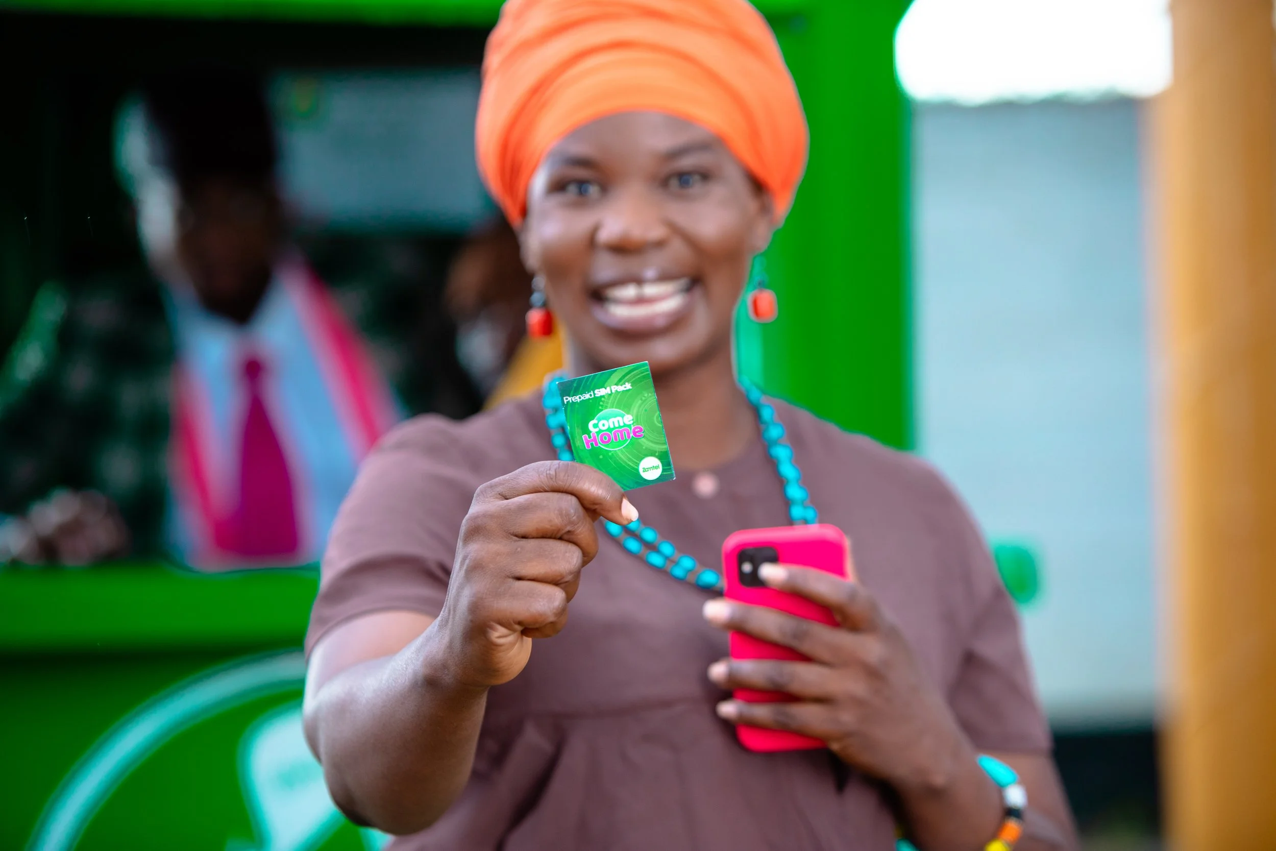 Woman wearing an orange headwrap and turquoise jewelry holding a green SIM card and a pink smartphone, smiling at the camera.