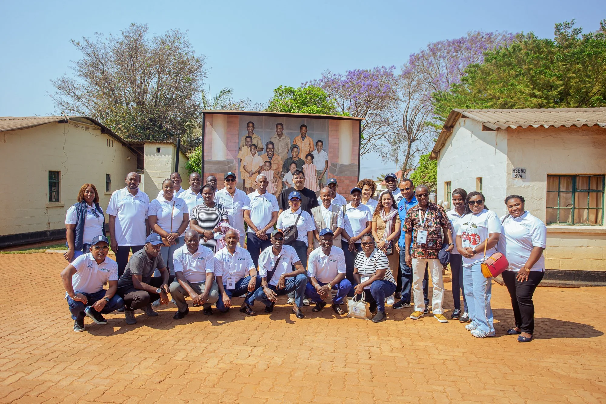 A large group of diverse people posing outdoors in front of a building and a mural depicting a family portrait, with trees and a clear sky in the background.