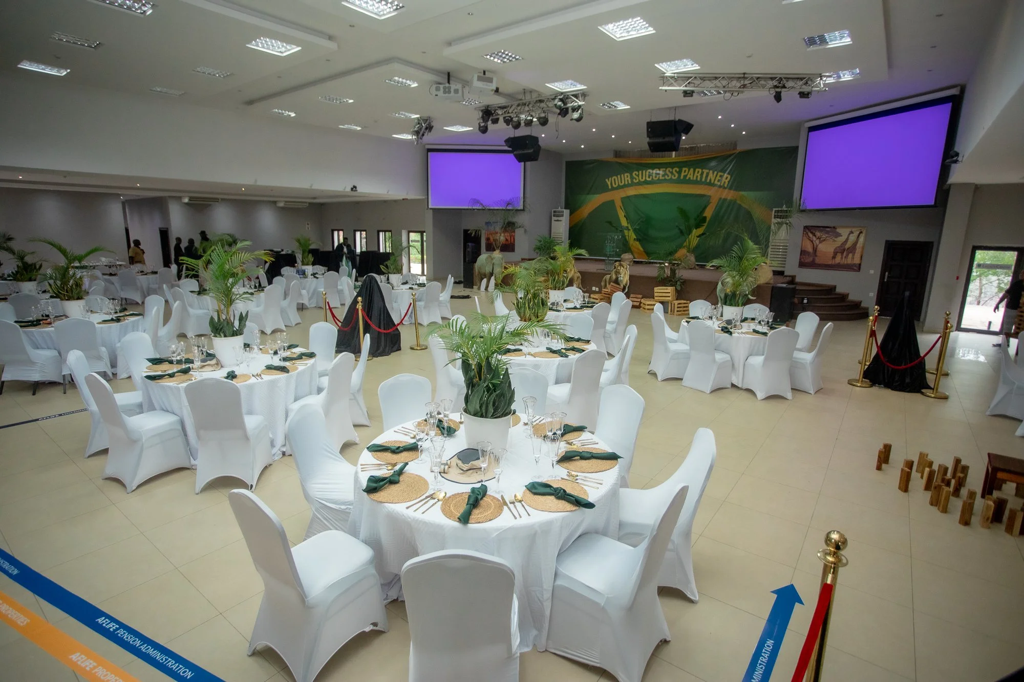 A large event hall decorated for a formal gathering or banquet, featuring round tables with white tablecloths, chairs covered in white fabric, and centerpieces with green plants. The stage at the front has two large screens with purple backgrounds an