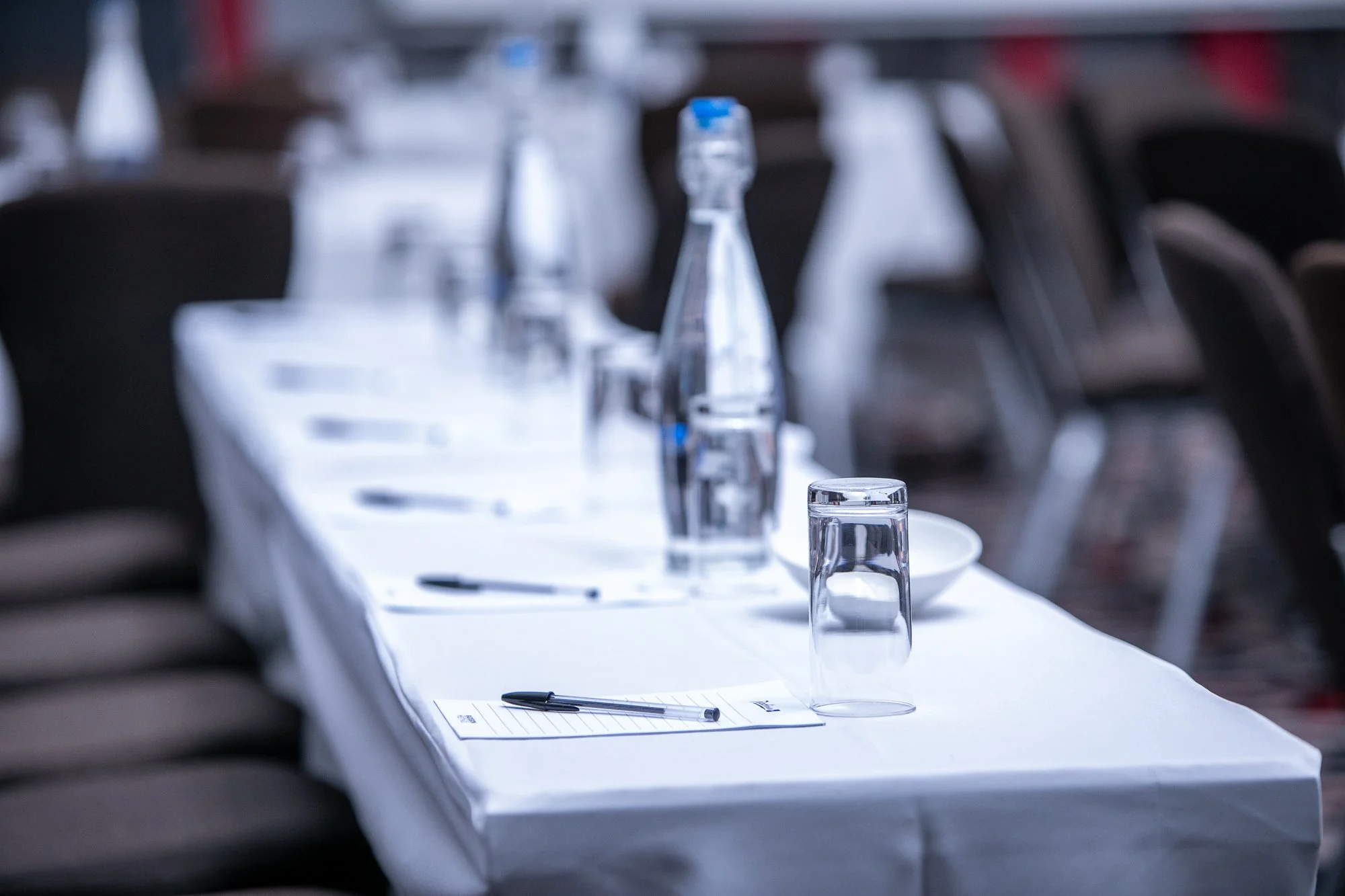 A conference room with a long table set for a meeting, with water bottles, glasses, notepads, and pens, surrounded by empty chairs.