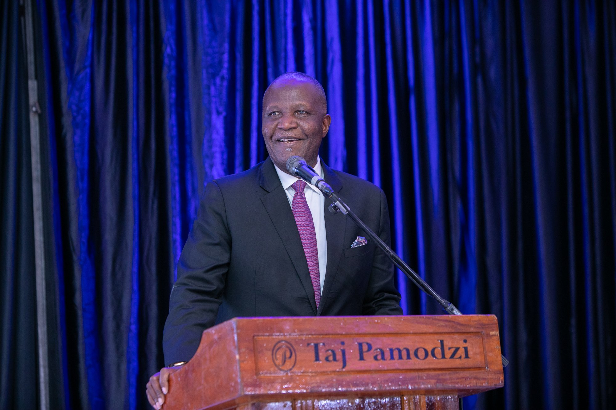 A man in a black suit with a pink tie speaking at a podium with a microphone, in front of dark blue curtains, at an event at Taj Pamozdi.
